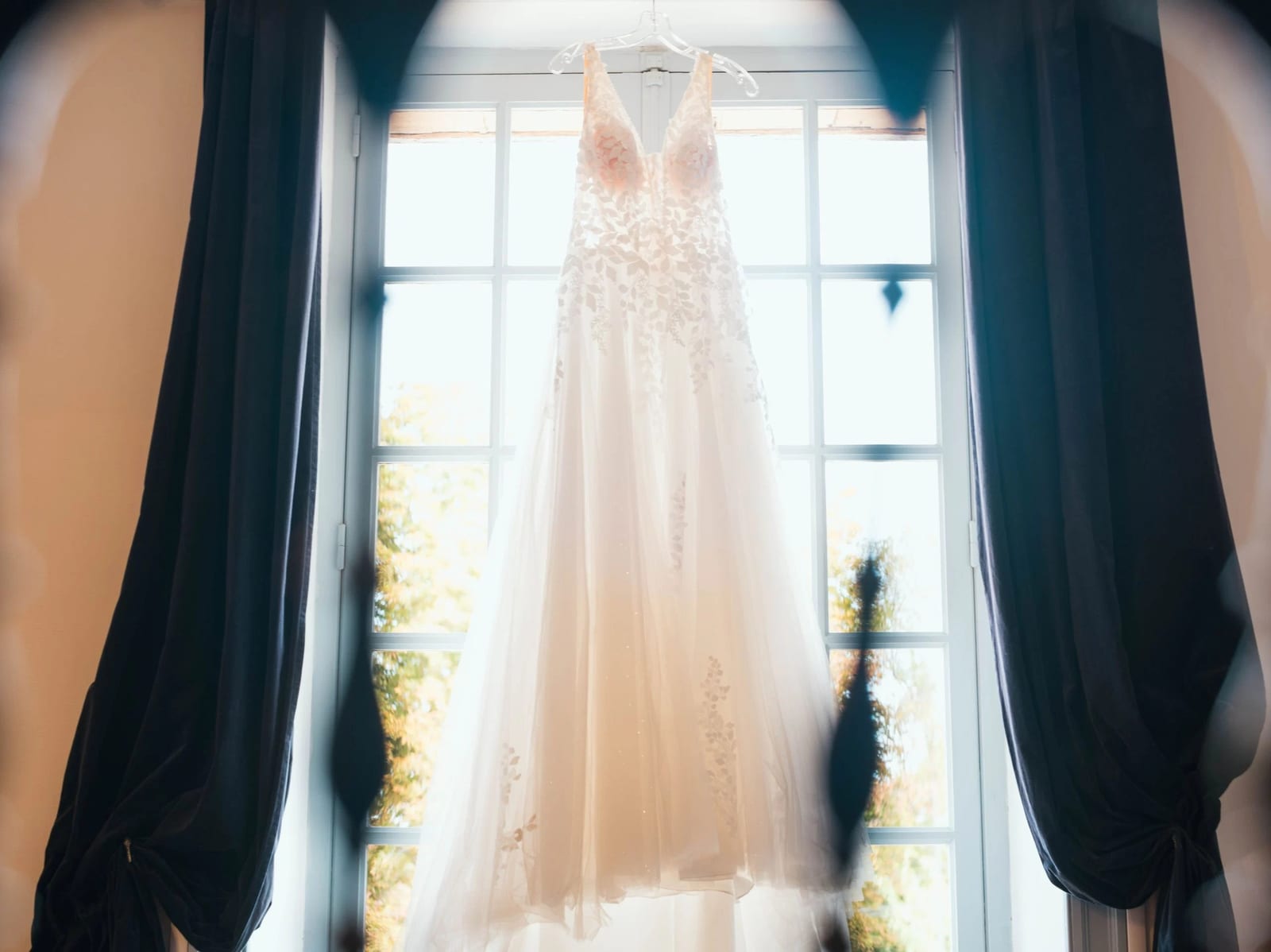 La robe de mariée d'Amélie, bustier de dentelle florale et traîne de tulle, suspendue devant la fenêtre de la chambre, mariage au Les Voûtes du Plessier, par Jean-Baptiste Argyre (Jaysphotography)