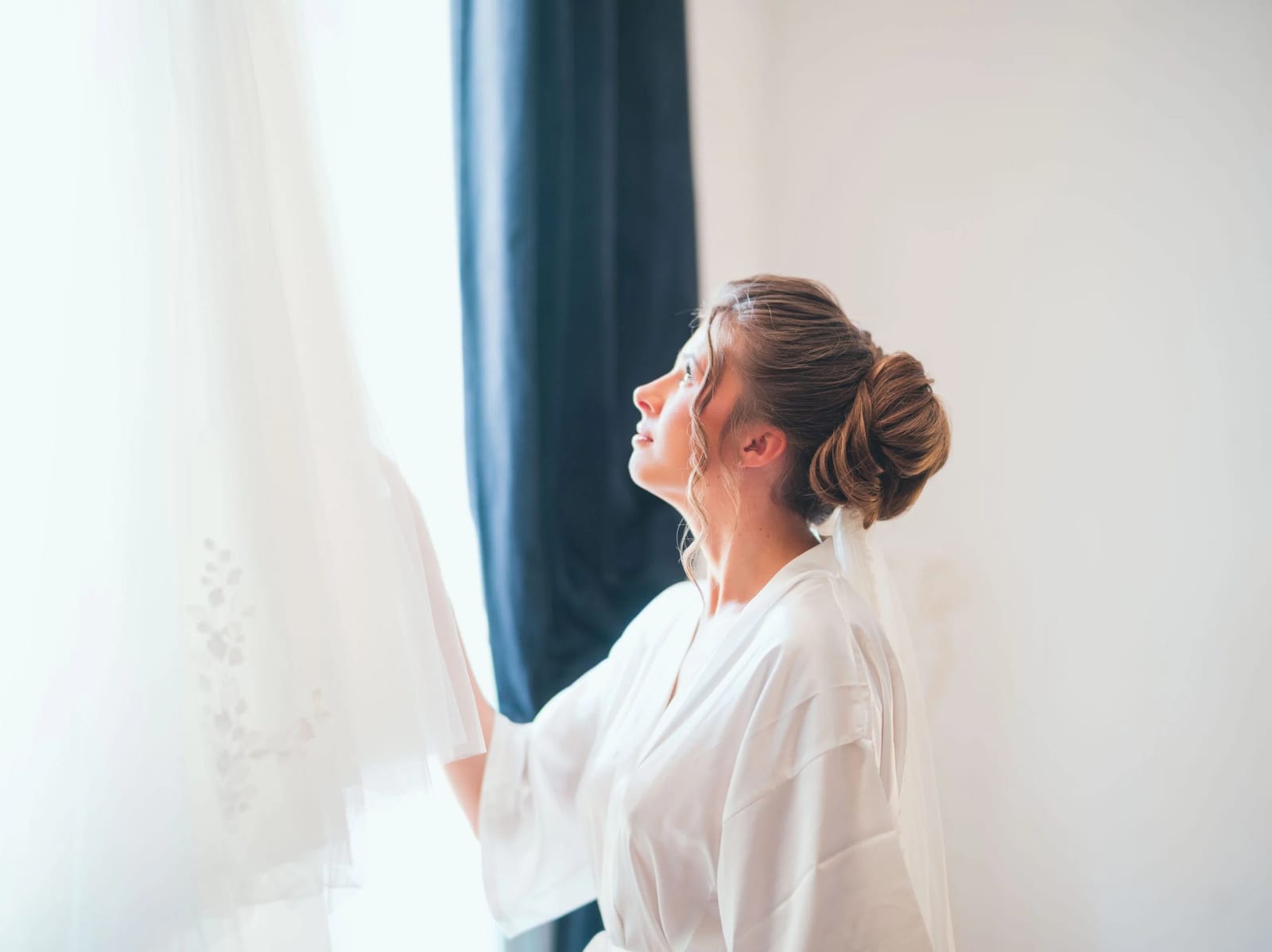 Amélie de profil en peignoir blanc devant le voilage de la fenêtre, chignon coiffé, avant l'habillage, mariage au Les Voûtes du Plessier, par Jean-Baptiste Argyre (Jaysphotography)