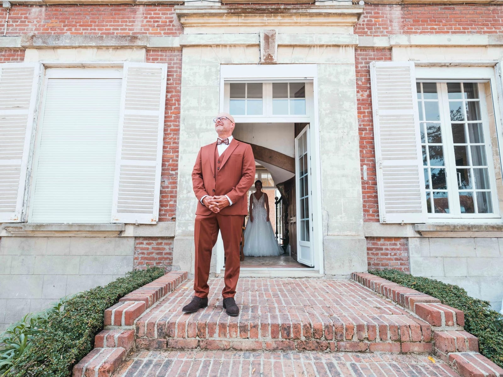 Le père d'Amélie en costume terracotta attend sur le perron de la maison de brique pendant que sa fille apparaît en robe dans l'embrasure, mariage au Les Voûtes du Plessier, par Jean-Baptiste Argyre (Jaysphotography)