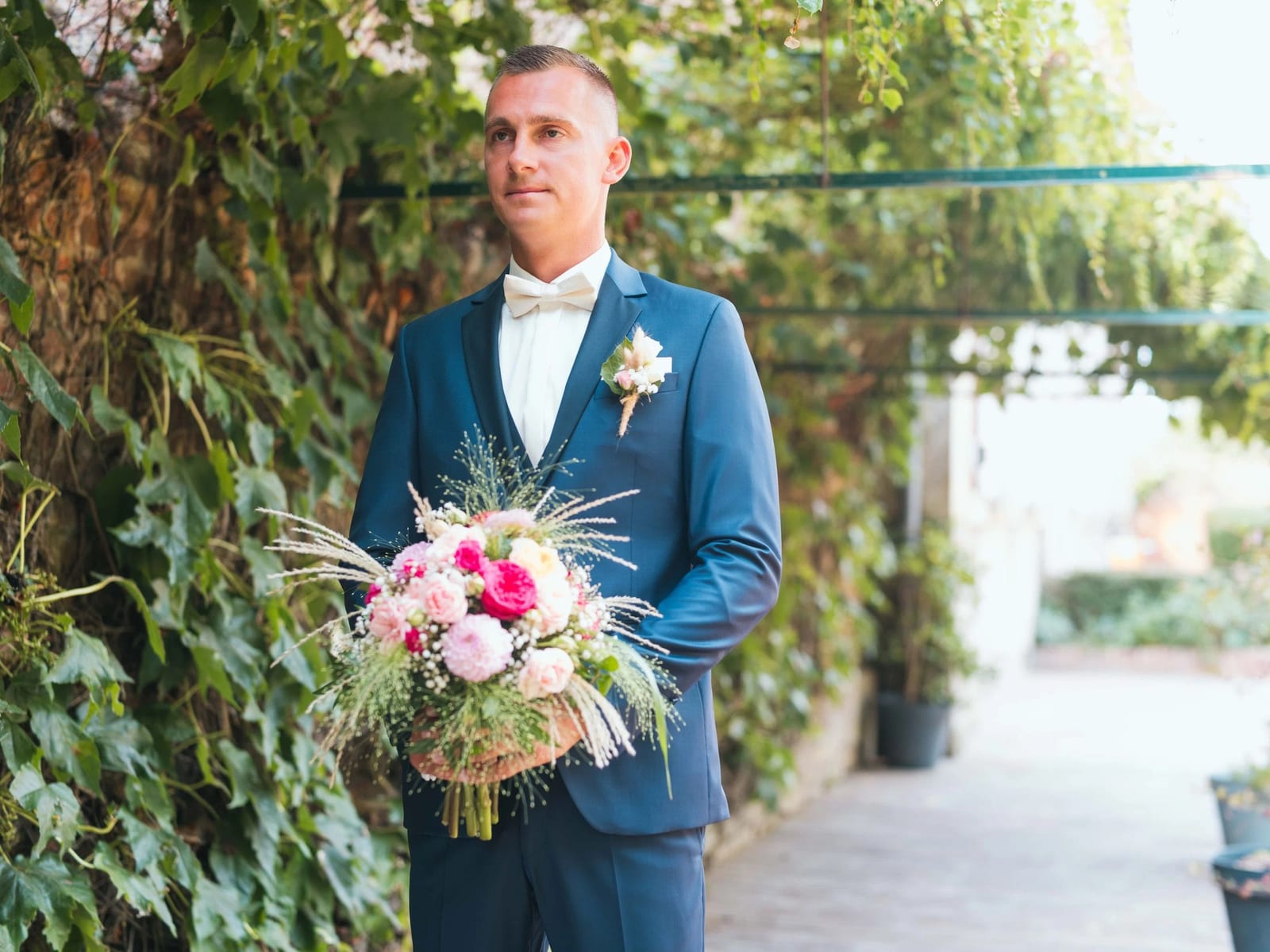 Guillaume en costume bleu attend sous la pergola de vigne vierge, le bouquet rose et fuchsia de la mariée entre les mains, mariage au Les Voûtes du Plessier, par Jean-Baptiste Argyre (Jaysphotography)