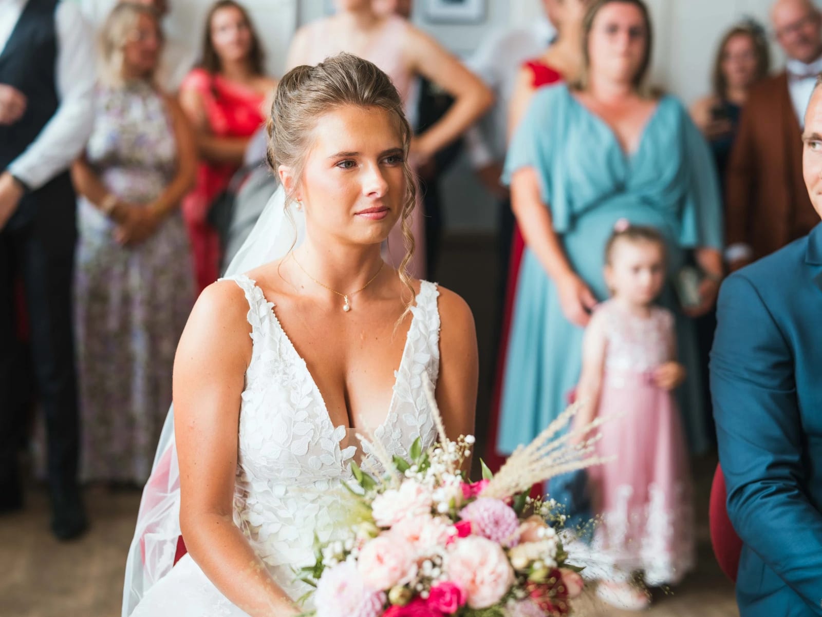 Amélie assise dans la salle des mariages, son bouquet sur les genoux, invités debout au fond, mariage au Les Voûtes du Plessier, par Jean-Baptiste Argyre (Jaysphotography)