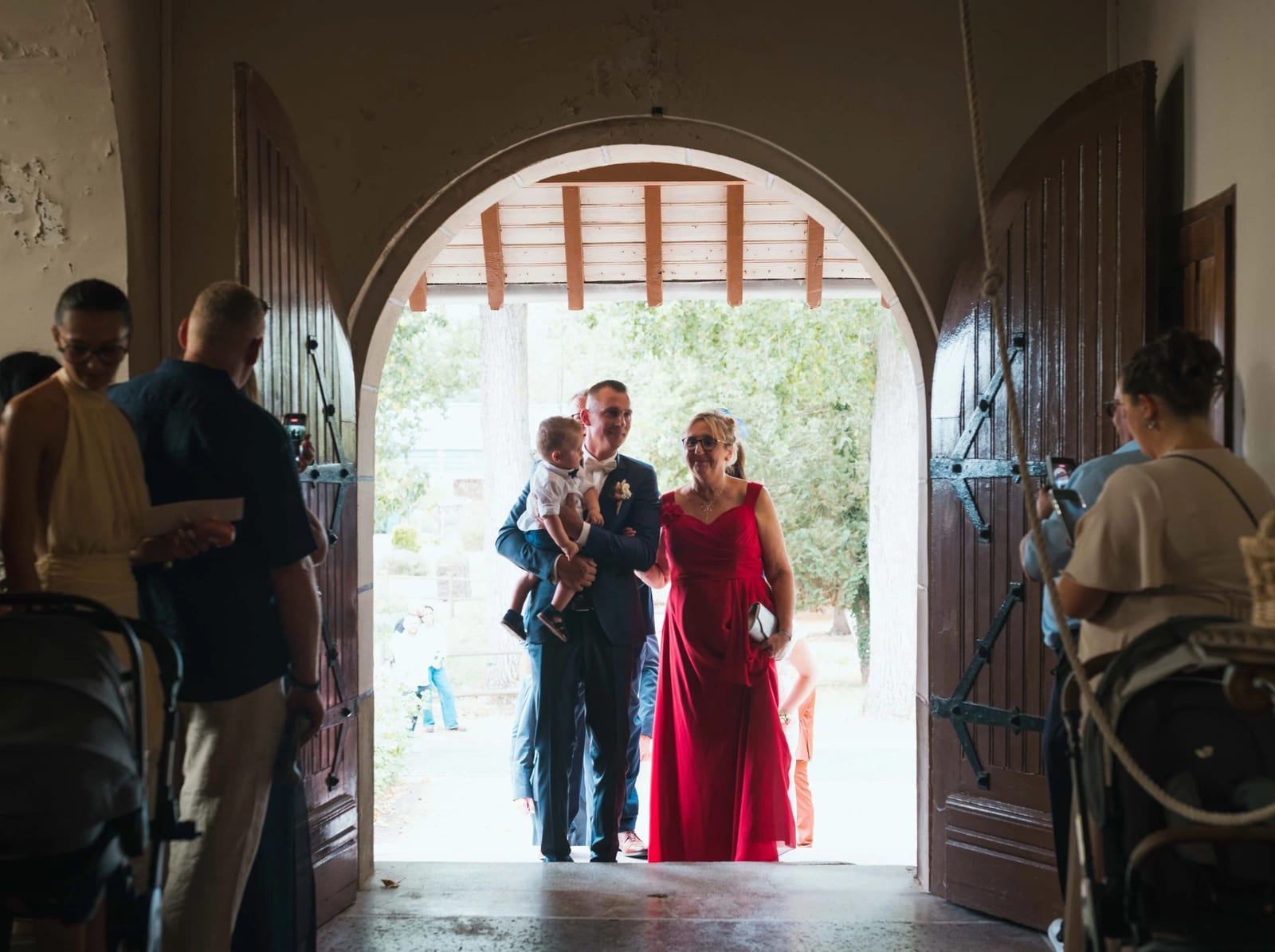 Entrée à l'église : Guillaume porte son fils et avance au bras de sa mère en robe rouge sous le porche ouvert, mariage au Les Voûtes du Plessier, par Jean-Baptiste Argyre (Jaysphotography)