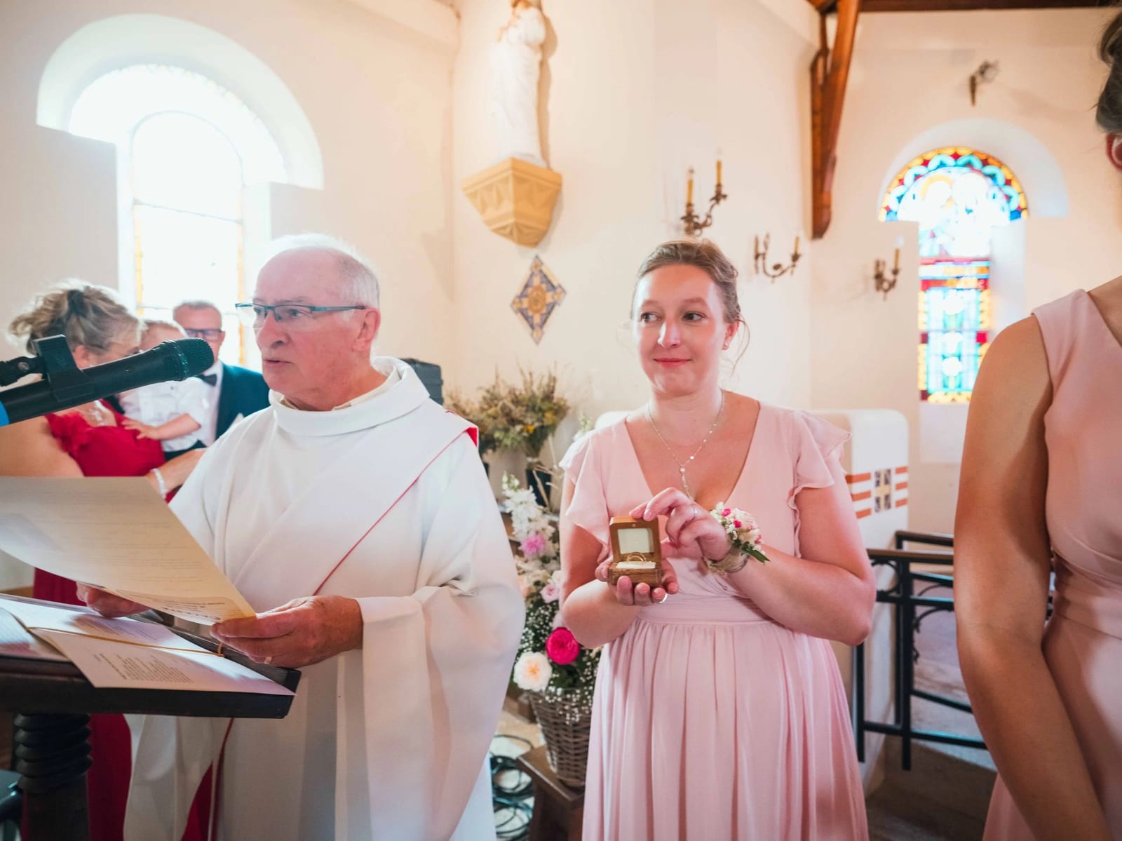 Une demoiselle d'honneur présente les alliances dans leur écrin devant le prêtre, à l'autel, mariage au Les Voûtes du Plessier, par Jean-Baptiste Argyre (Jaysphotography)
