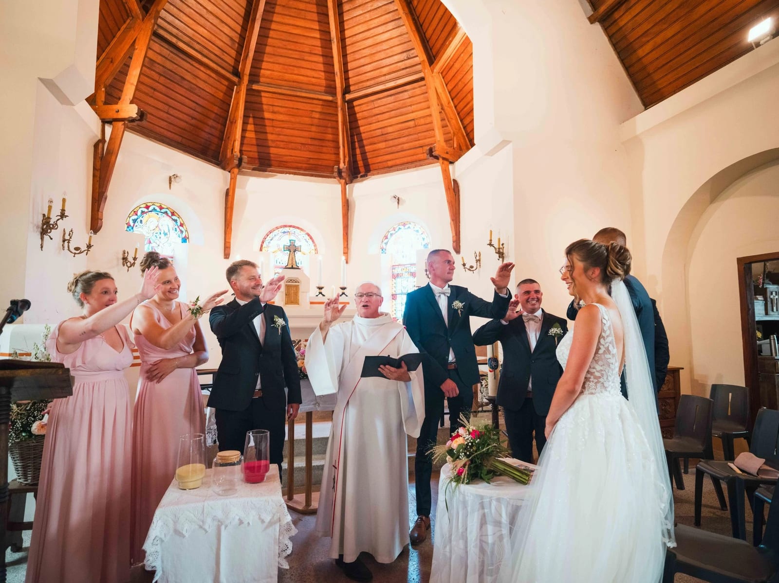 Bénédiction des mariés : les proches tendent la main au-dessus d'Amélie et Guillaume, sous la charpente en éventail de l'église, mariage au Les Voûtes du Plessier, par Jean-Baptiste Argyre (Jaysphotography)