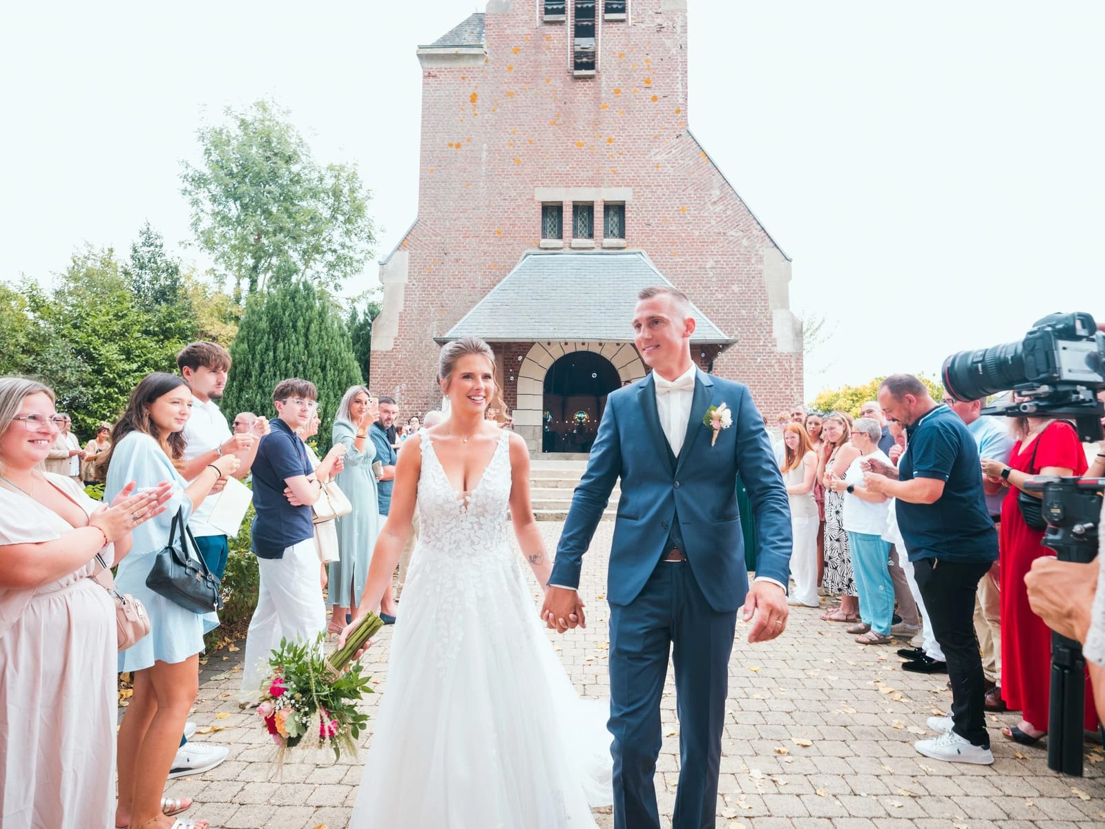 Sortie d'église main dans la main devant le clocher de brique, les invités faisant la haie, mariage au Les Voûtes du Plessier, par Jean-Baptiste Argyre (Jaysphotography)