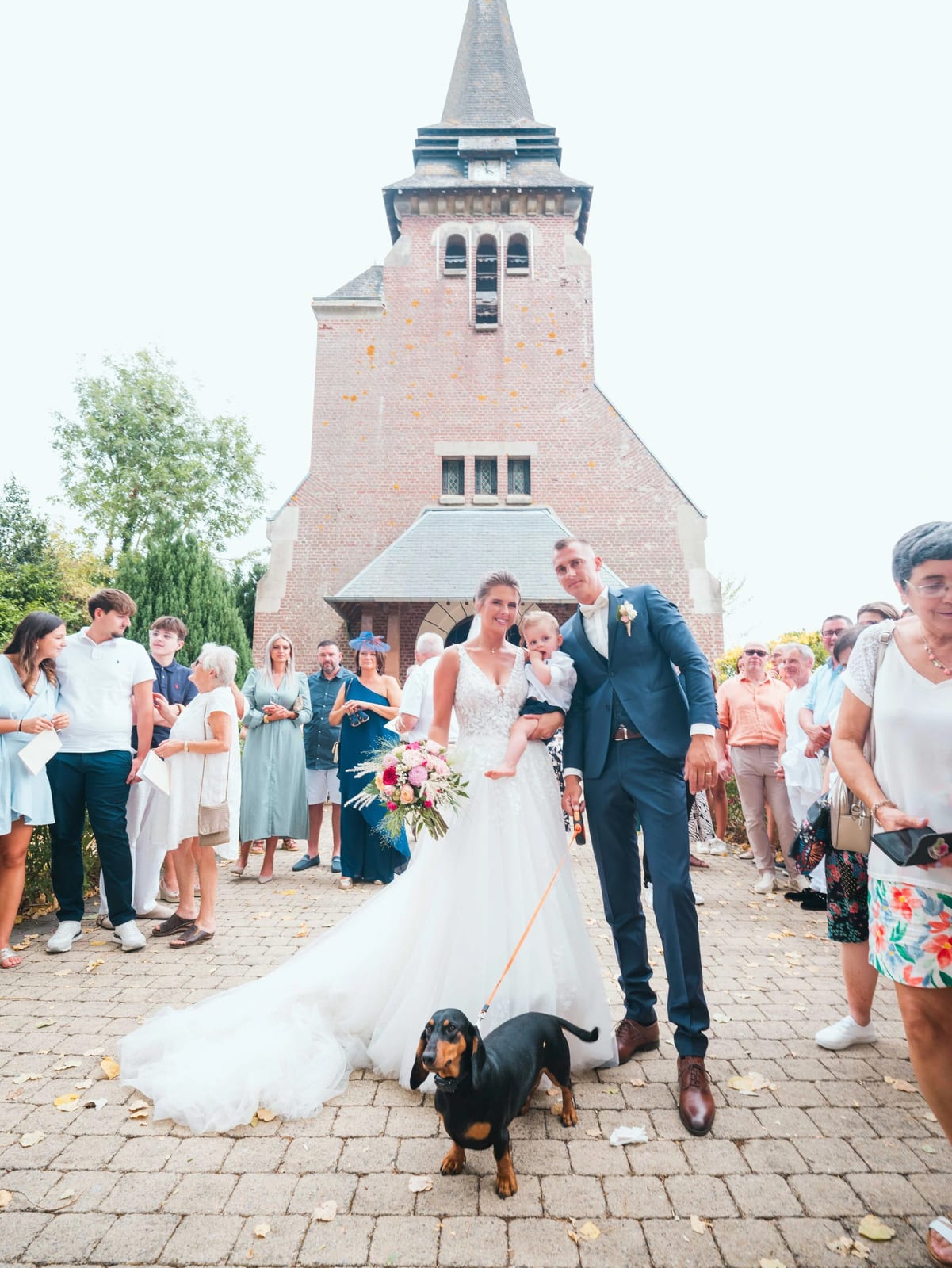 Le teckel du couple, en laisse orange, planté devant Amélie et Guillaume à la sortie de l'église, mariage au Les Voûtes du Plessier, par Jean-Baptiste Argyre (Jaysphotography)