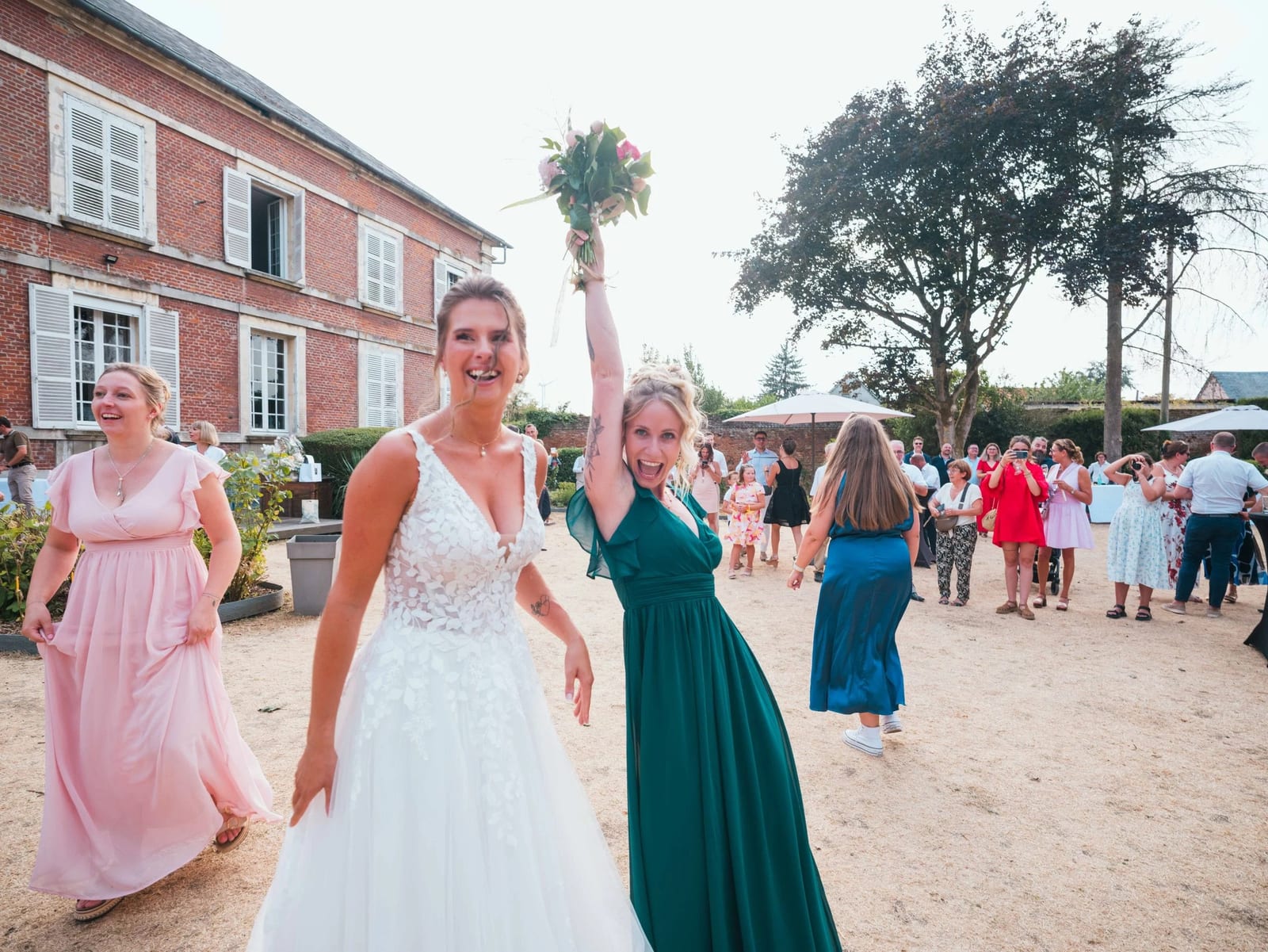 Lancer de bouquet : une demoiselle d'honneur en vert bouteille l'attrape, les deux bras levés, au milieu des invitées, mariage au Les Voûtes du Plessier, par Jean-Baptiste Argyre (Jaysphotography)