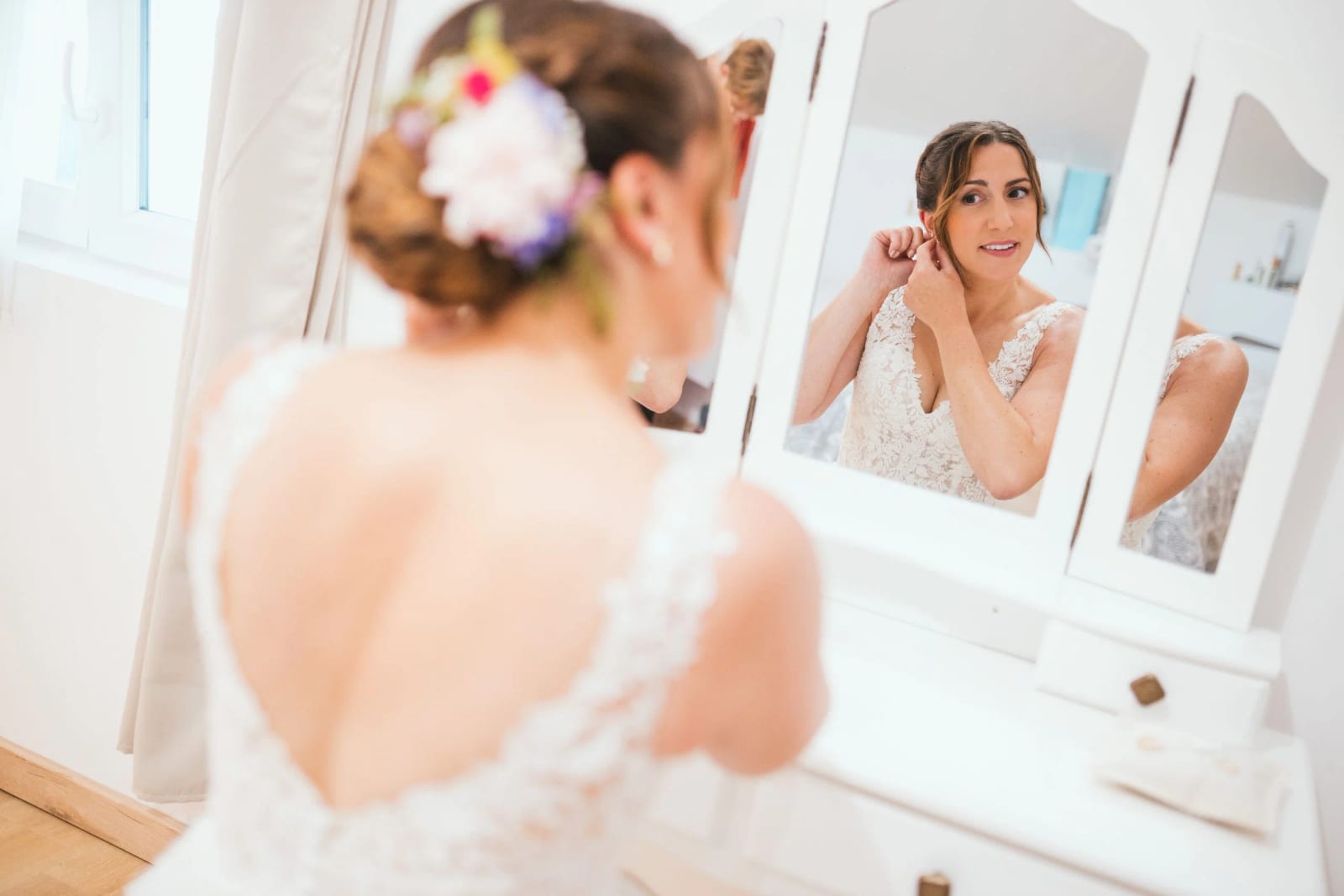La mariée de dos devant un miroir à trois pans, dos de robe en dentelle ajourée et fleurs dans les cheveux, La Mézandrée, mariage au La Mézandrée, par Jean-Baptiste Argyre (Jaysphotography)