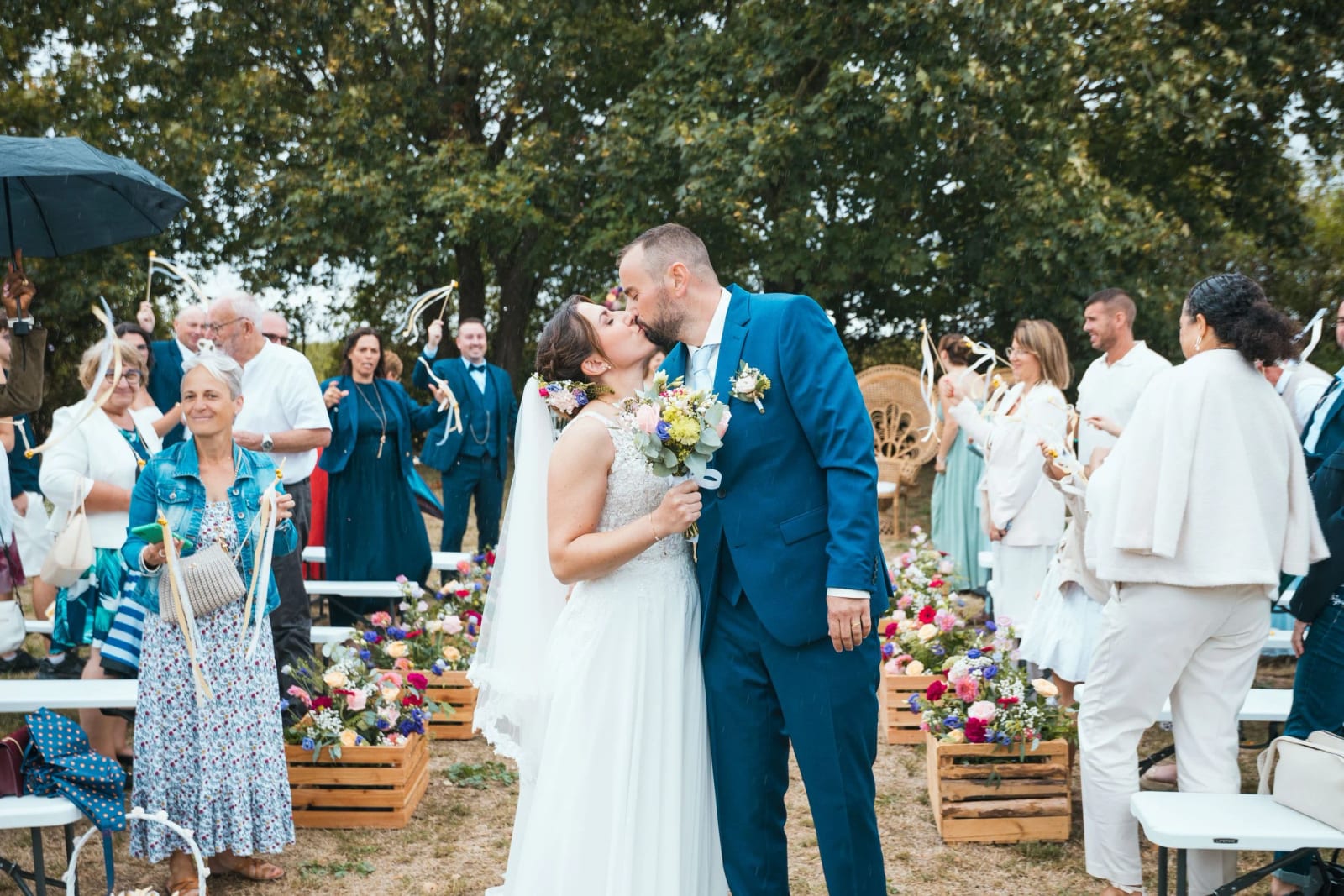 Sortie de cérémonie dans l'allée bordée de cagettes fleuries, les invités agitent des bâtons à rubans, La Mézandrée à Prouvais, mariage au La Mézandrée, par Jean-Baptiste Argyre (Jaysphotography)