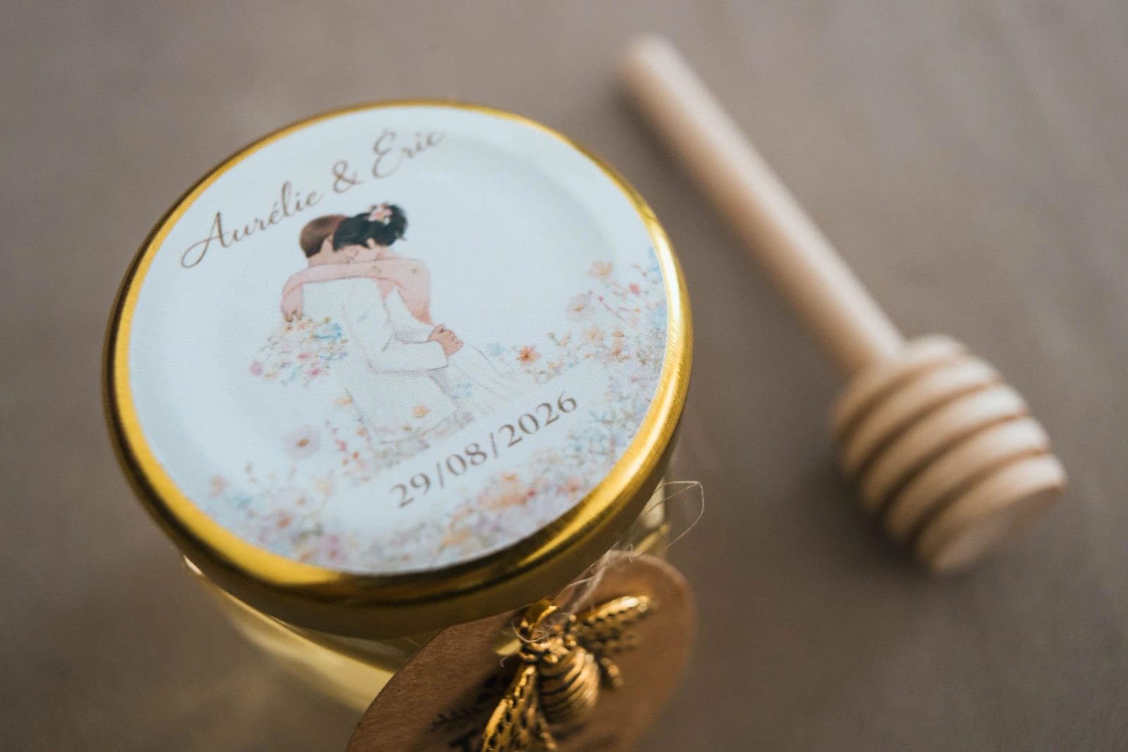 Le cadeau d'invités : pot de miel à couvercle doré illustré du couple, prénoms et date du 29 août 2026, abeille en breloque et cuillère à miel, mariage au La Mézandrée, par Jean-Baptiste Argyre (Jaysphotography)