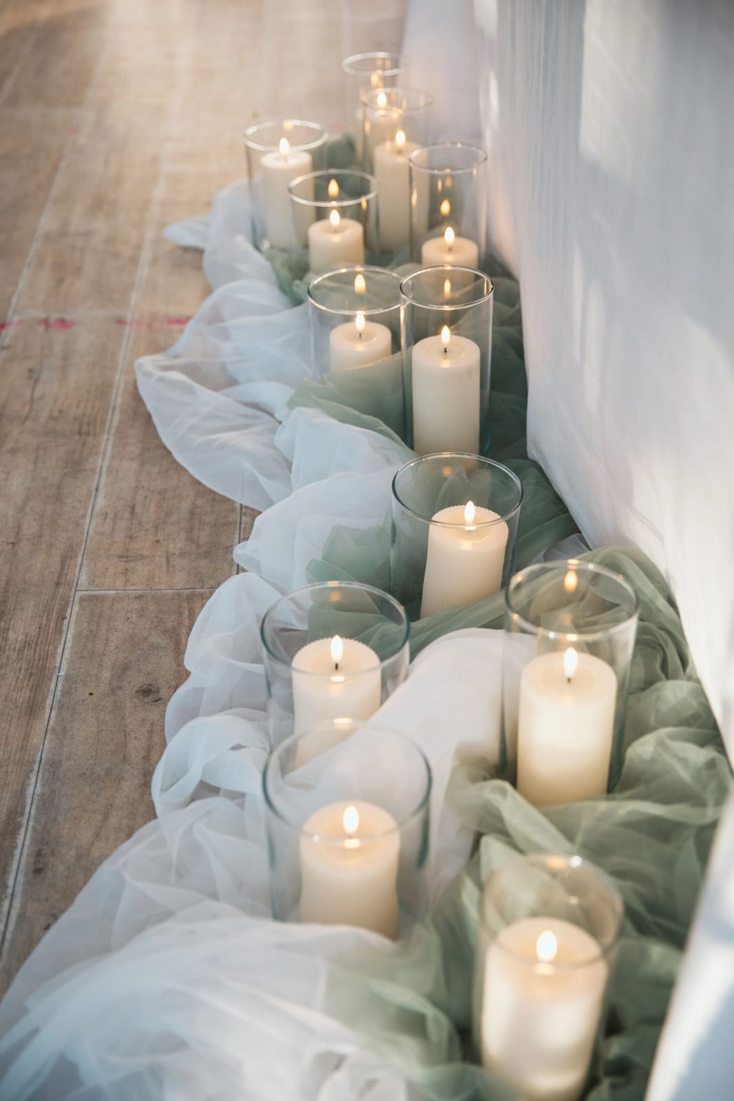 Rangée de bougies dans des photophores de verre sur un chemin de tulle blanc et vert sauge, le long d'un mur drapé de blanc, mariage au Château de la Houssoye, par Jean-Baptiste Argyre (Jaysphotography)