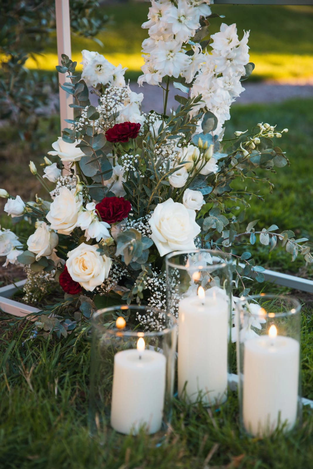 Composition de delphiniums et roses blanches, roses rouges, gypsophile et eucalyptus avec trois bougies sur l'herbe, mariage au Château de la Houssoye, par Jean-Baptiste Argyre (Jaysphotography)
