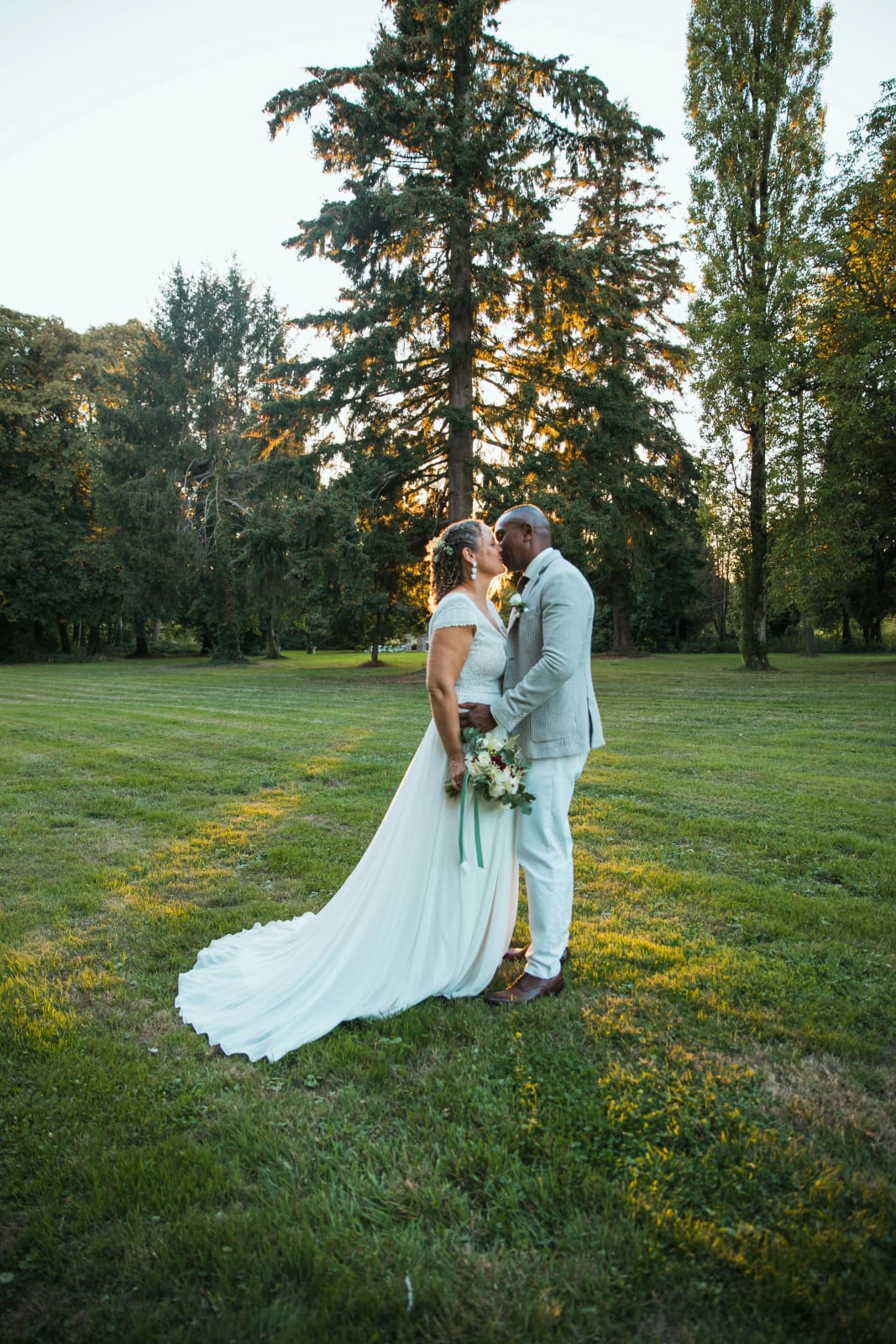 Les mariés s'embrassent au milieu d'une grande pelouse bordée de hauts conifères et de peupliers, traîne étalée dans l'herbe, mariage au Château de la Houssoye, par Jean-Baptiste Argyre (Jaysphotography)