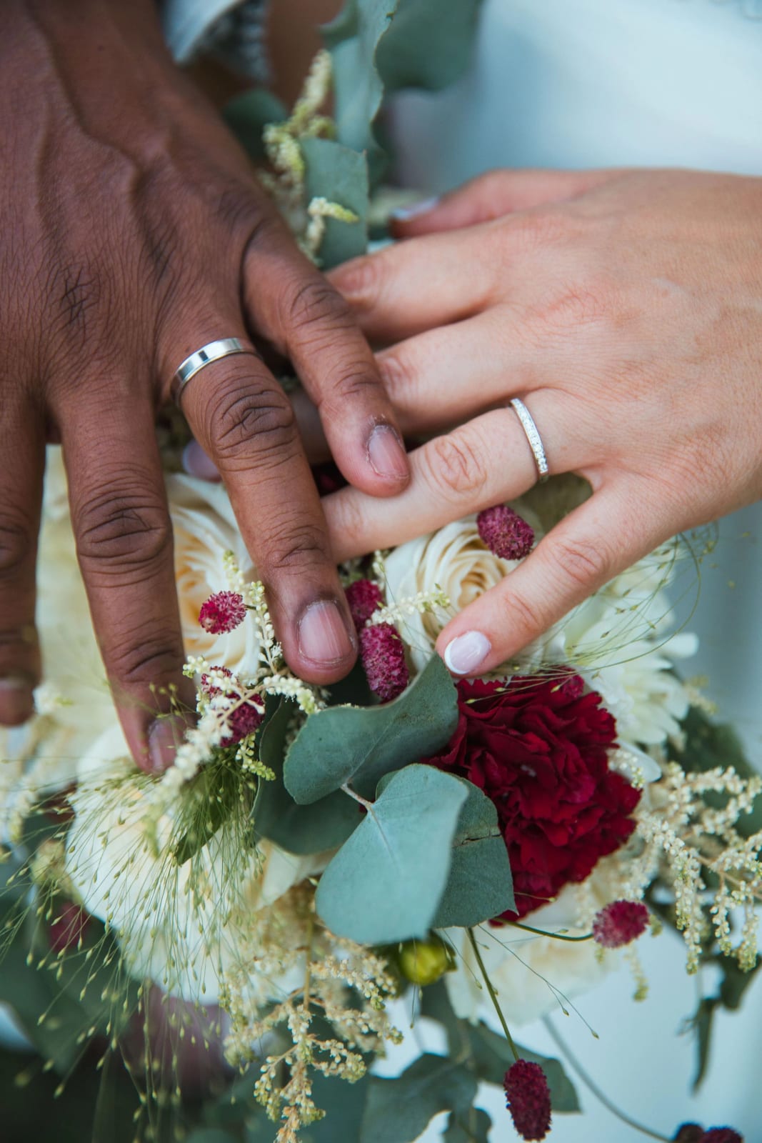 Les mains des mariés avec leurs alliances posées sur le bouquet de roses blanches, rose rouge et eucalyptus, mariage au Château de la Houssoye, par Jean-Baptiste Argyre (Jaysphotography)