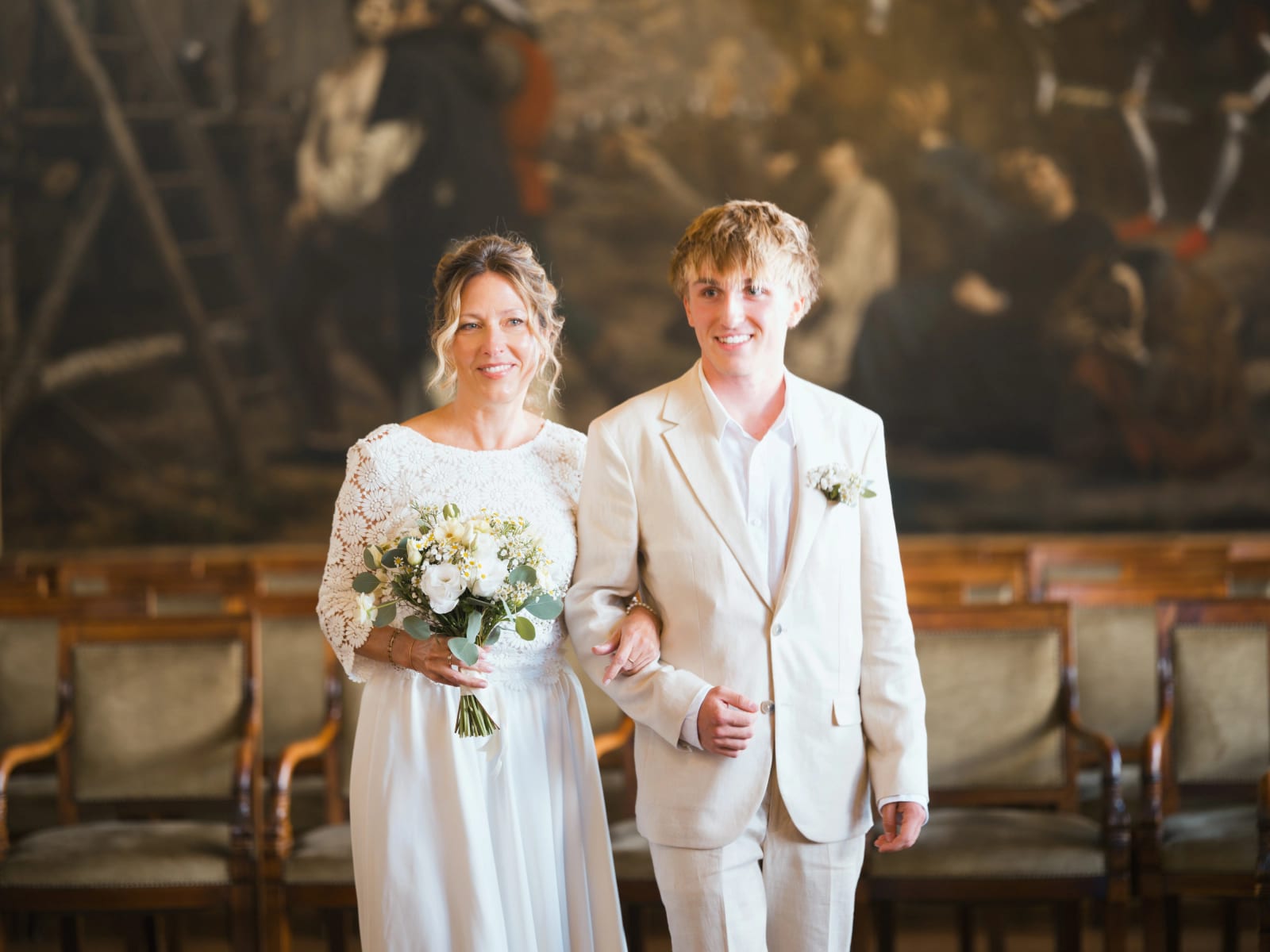 La mariée en robe à haut de dentelle fleurie entre dans la salle des mariages de la mairie de Senlis au bras d'un jeune homme en costume écru, grande toile peinte au fond, mariage à la Mairie de Senlis, par Jean-Baptiste Argyre (Jaysphotography)