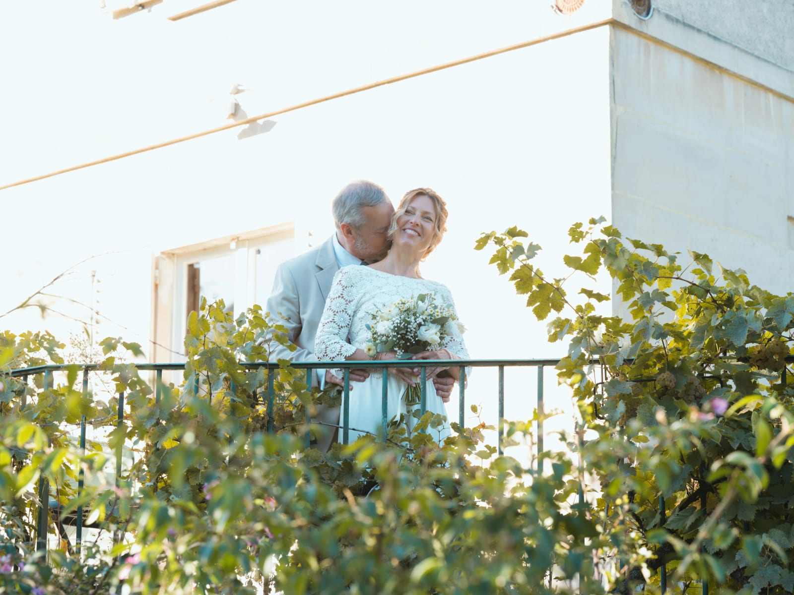 Le marié embrasse la mariée dans le cou sur un balcon à garde-corps vert couvert de vigne, mariage à la Mairie de Senlis, par Jean-Baptiste Argyre (Jaysphotography)
