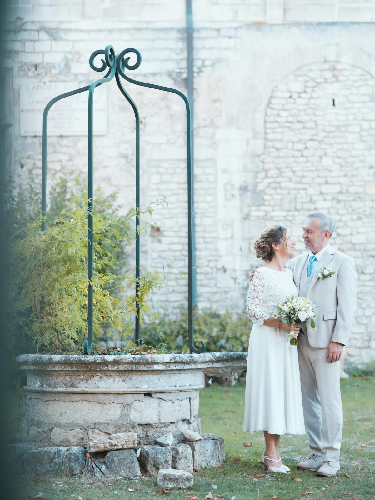 Les mariés près d'un ancien puits de pierre à armature de fer forgé vert, contre un haut mur de pierre, mariage à la Mairie de Senlis, par Jean-Baptiste Argyre (Jaysphotography)