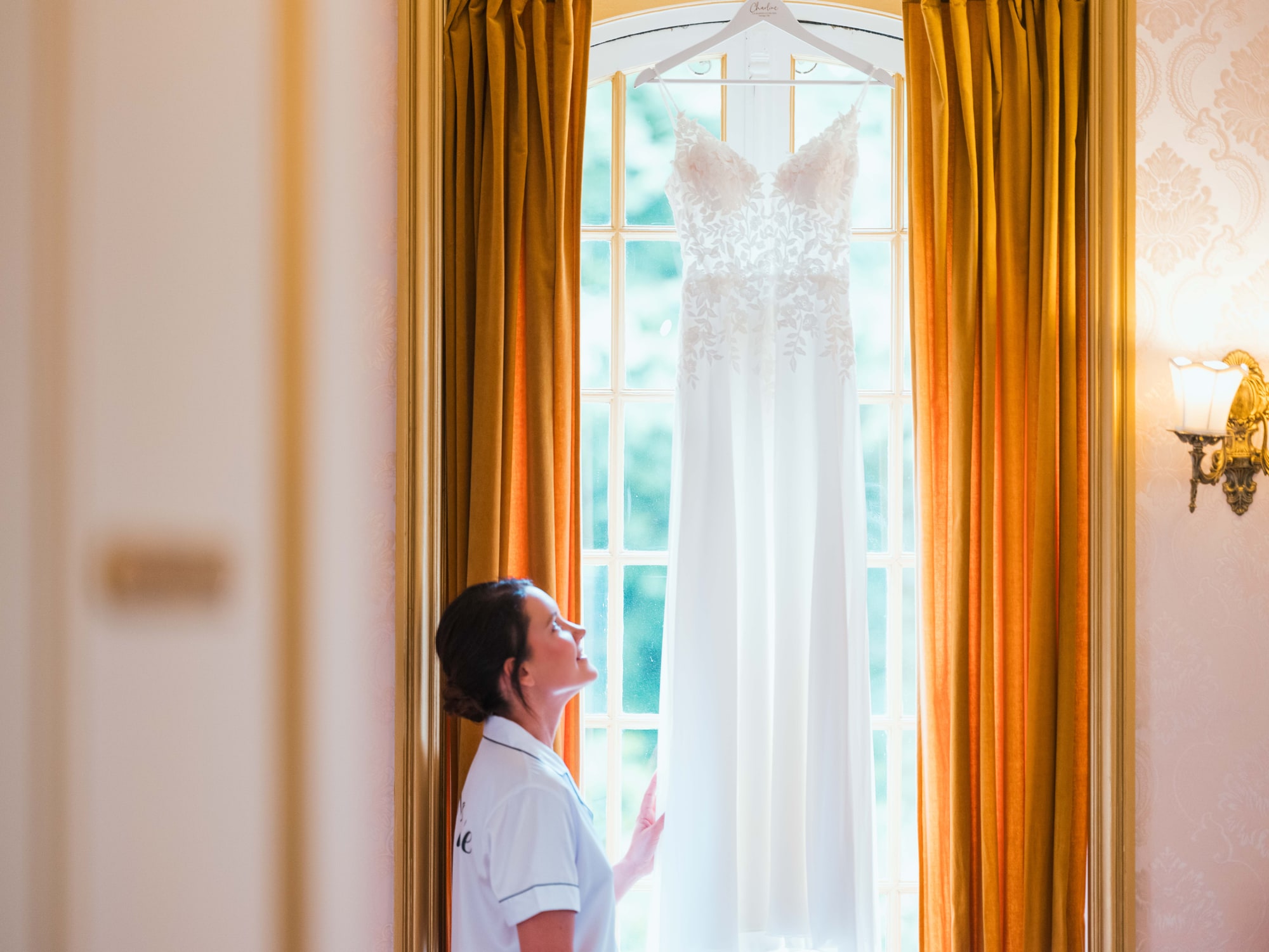 Charline en pyjama contemplant sa robe de mariée à la fenêtre dorée, mariage au Château de Moliens, par Jean-Baptiste Argyre (Jaysphotography)