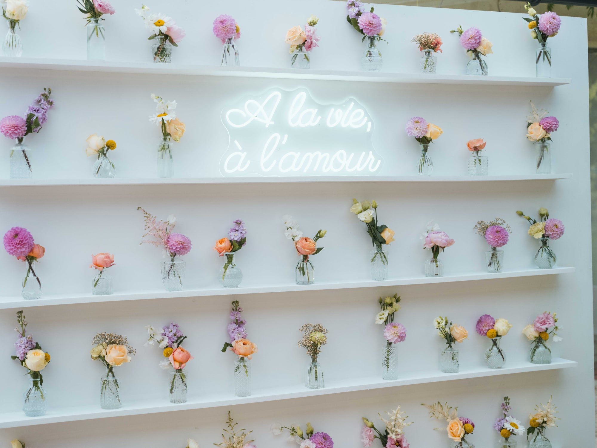 Mur de fleurs « À la vie, à l'amour » aux fioles pastel du vin d'honneur, mariage au Château de Moliens, par Jean-Baptiste Argyre (Jaysphotography)