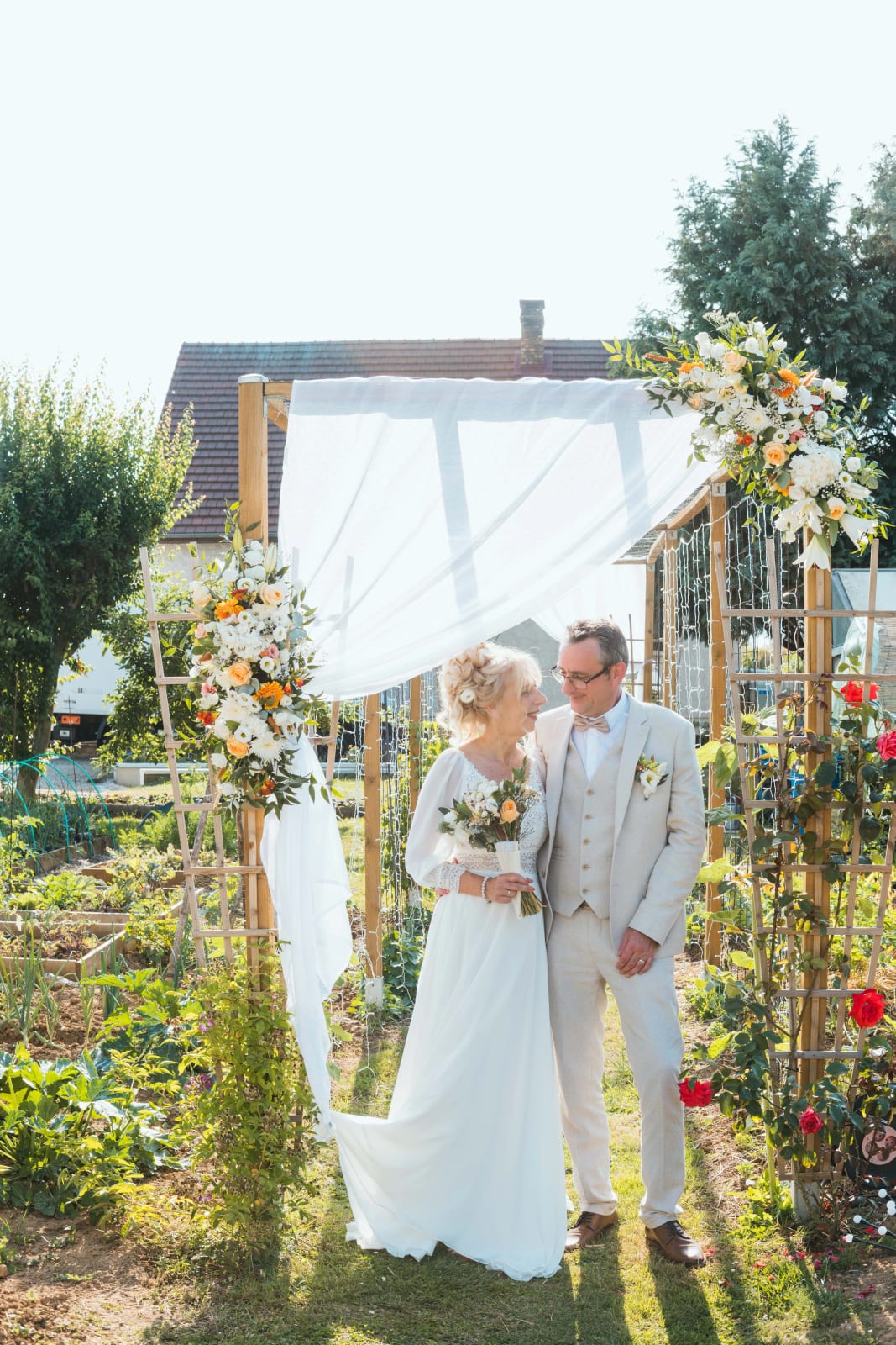 Corinne et Olivier sous l'arche fleurie du vin d'honneur à la maison, à Valescourt, mariage au Valescourt (Oise), par Jean-Baptiste Argyre (Jaysphotography)