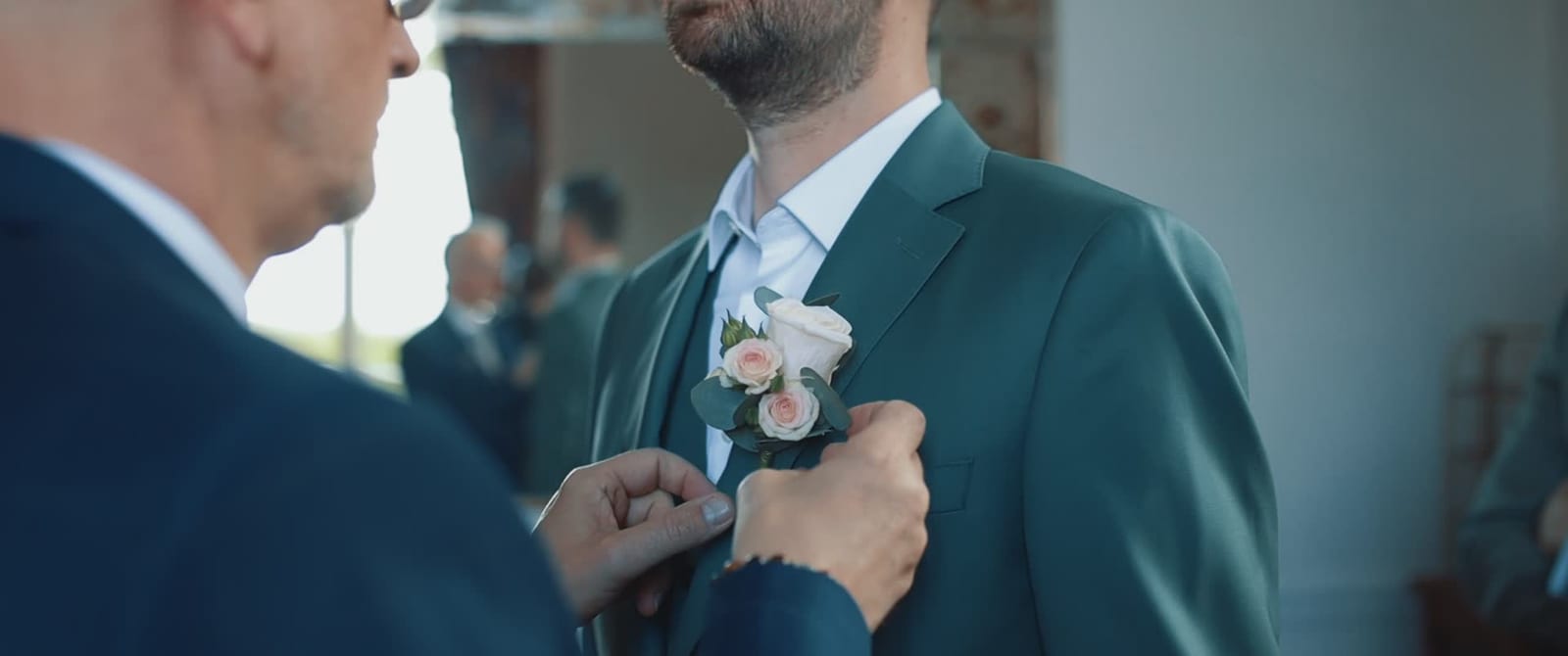 Un proche fixe la boutonnière de roses au costume vert de Thomas pendant les préparatifs, mariage au Château de Santeny, par Jean-Baptiste Argyre (Jaysphotography)