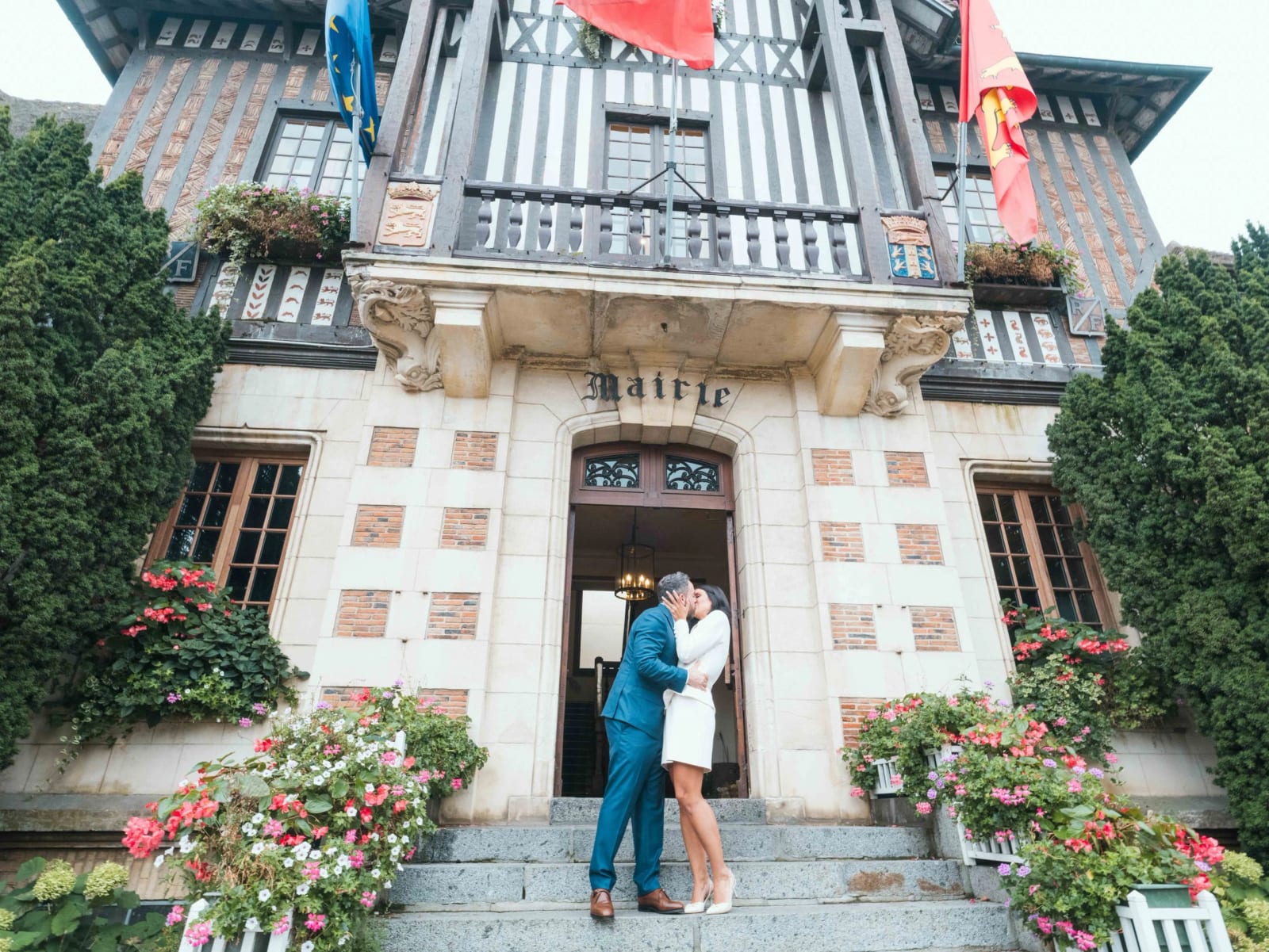 Les mariés s'embrassent sur les marches de la mairie de Deauville, façade à colombages et jardinières fleuries, mariage au Pavillon des Bains, Deauville, par Jean-Baptiste Argyre (Jaysphotography)
