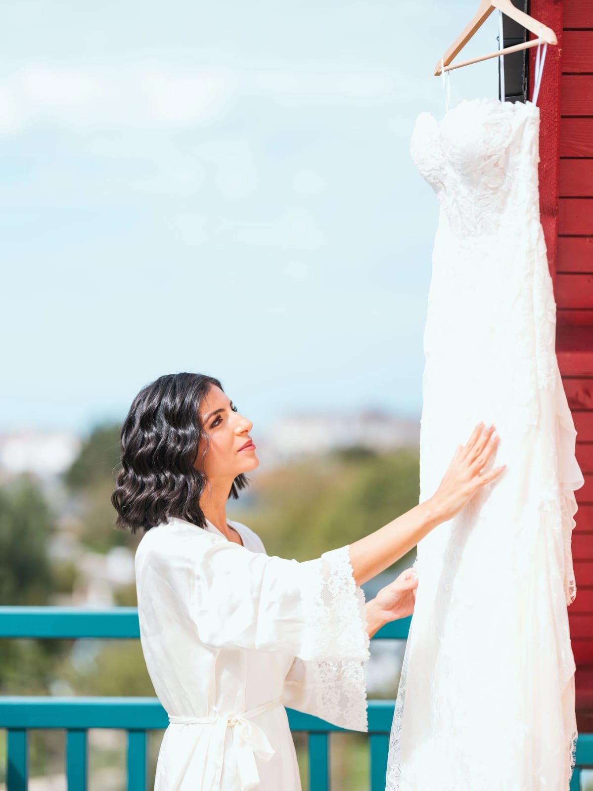 La mariée en peignoir de satin touche sa robe de dentelle suspendue contre un bardage rouge, balcon turquoise, mariage au Pavillon des Bains, Deauville, par Jean-Baptiste Argyre (Jaysphotography)