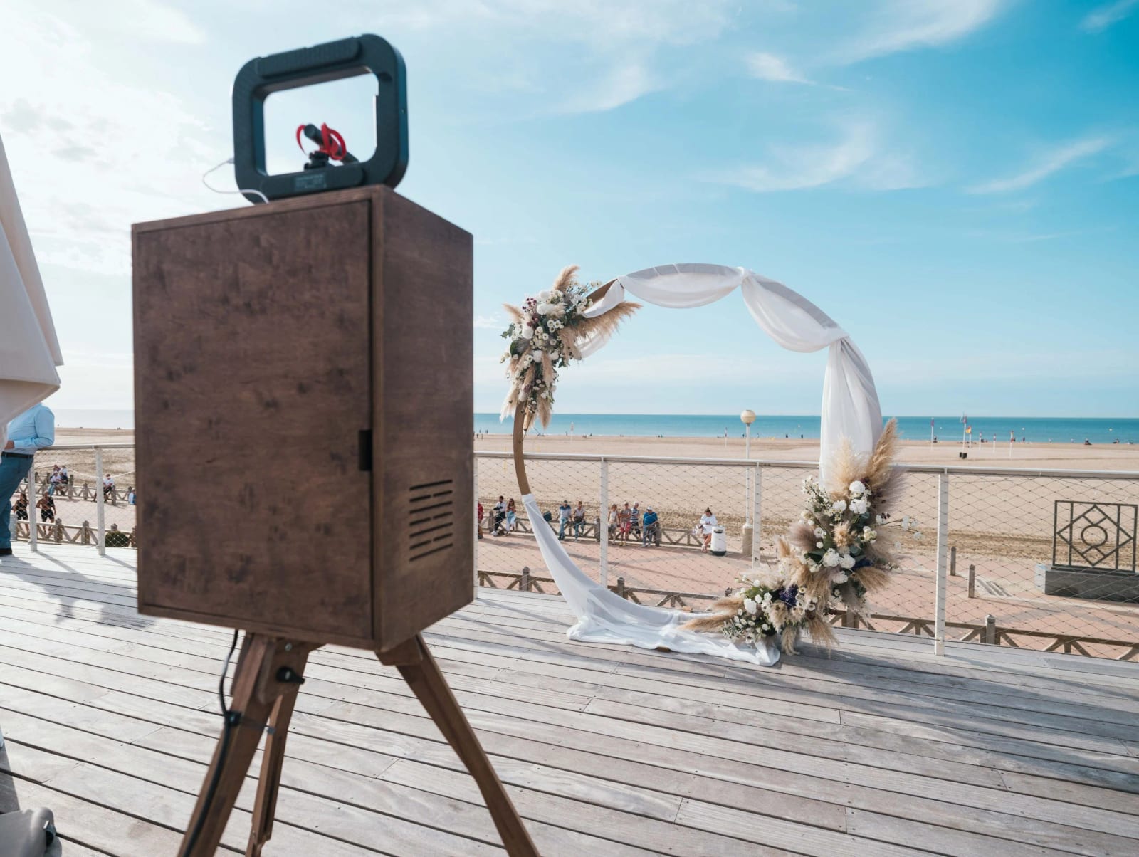 Arche ronde drapée de voile blanc et garnie de pampas sur une terrasse en bois face à la plage de Deauville, borne photo au premier plan, mariage au Pavillon des Bains, Deauville, par Jean-Baptiste Argyre (Jaysphotography)