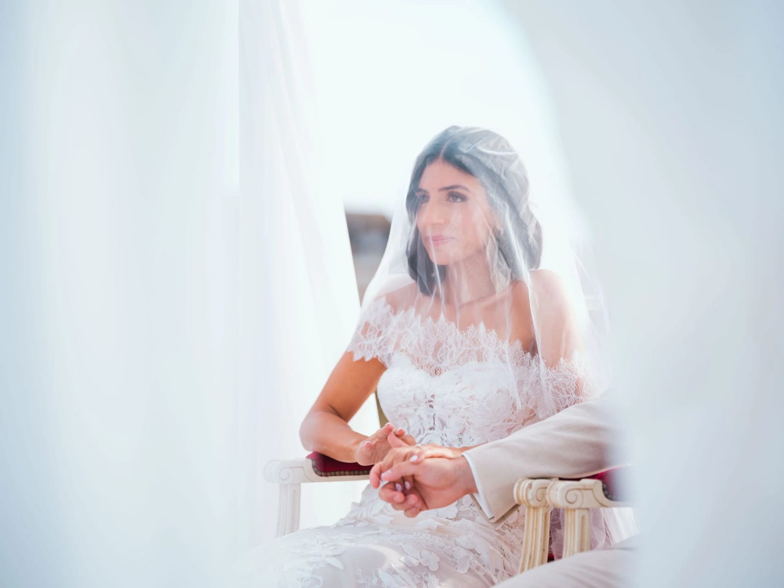 La mariée voilée assise dans un fauteuil blanc tient la main du marié pendant la cérémonie, à travers un voilage, mariage au Pavillon des Bains, Deauville, par Jean-Baptiste Argyre (Jaysphotography)