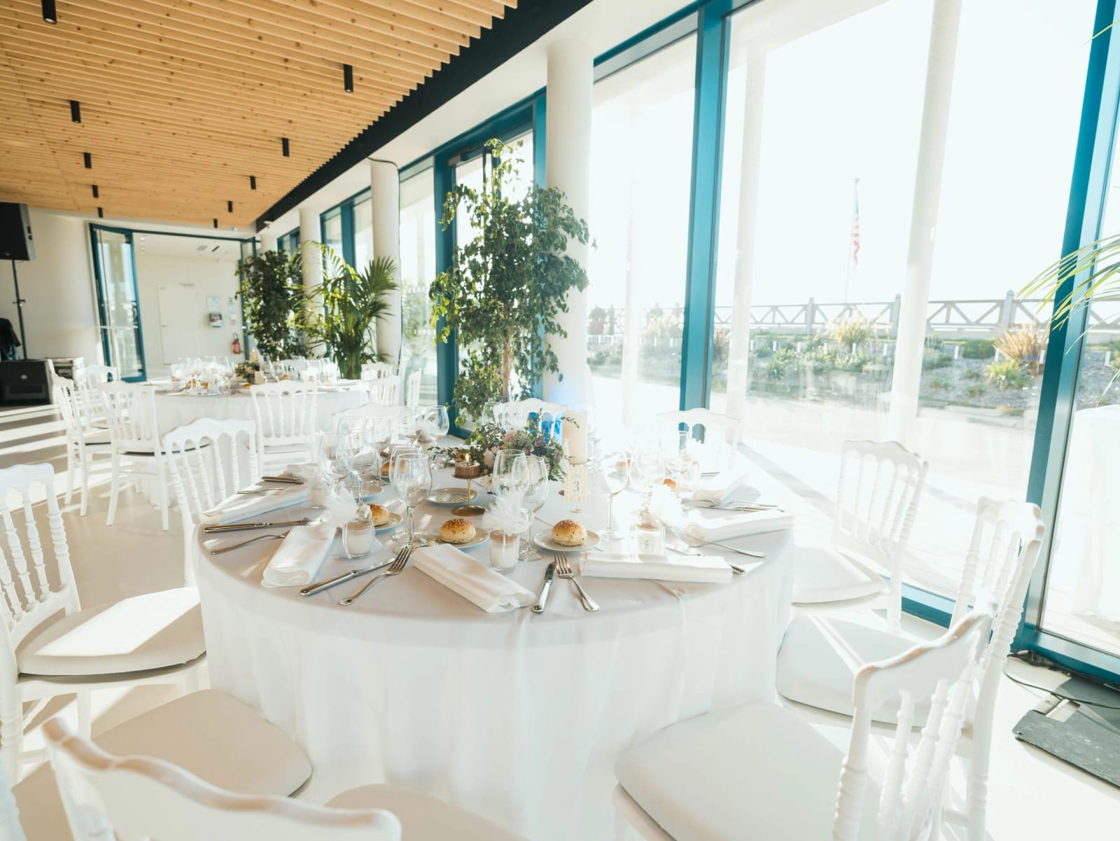 Salle de réception dressée : tables rondes blanches, chaises Napoléon III, baies vitrées cerclées de bleu sur le front de mer, mariage au Pavillon des Bains, Deauville, par Jean-Baptiste Argyre (Jaysphotography)