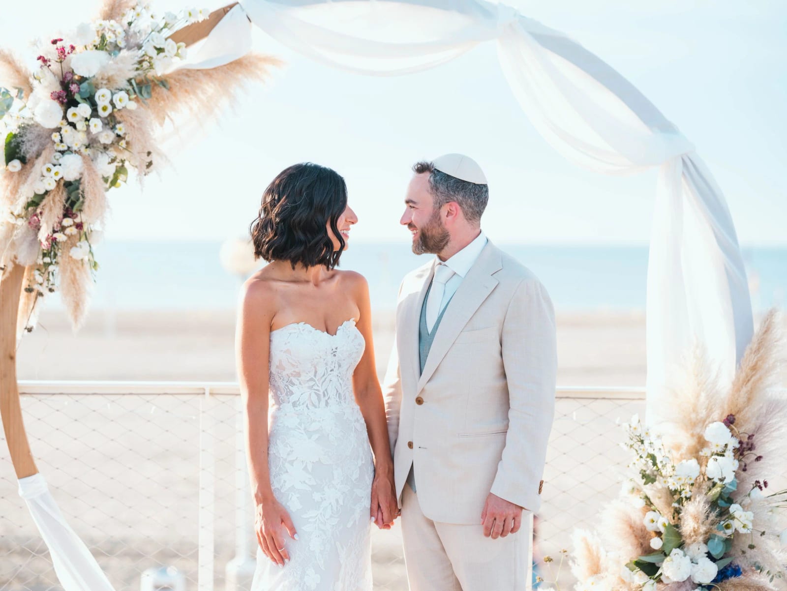 Les mariés face à face sous l'arche fleurie de pampas, la mer de Deauville en fond, mariage au Pavillon des Bains, Deauville, par Jean-Baptiste Argyre (Jaysphotography)