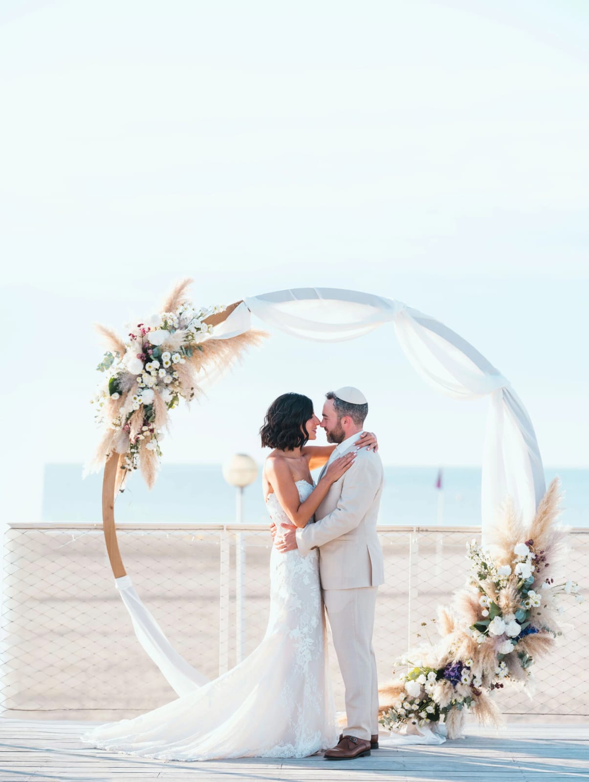 Les mariés enlacés sous l'arche ronde, traîne de dentelle sur la terrasse en bois, mariage au Pavillon des Bains, Deauville, par Jean-Baptiste Argyre (Jaysphotography)