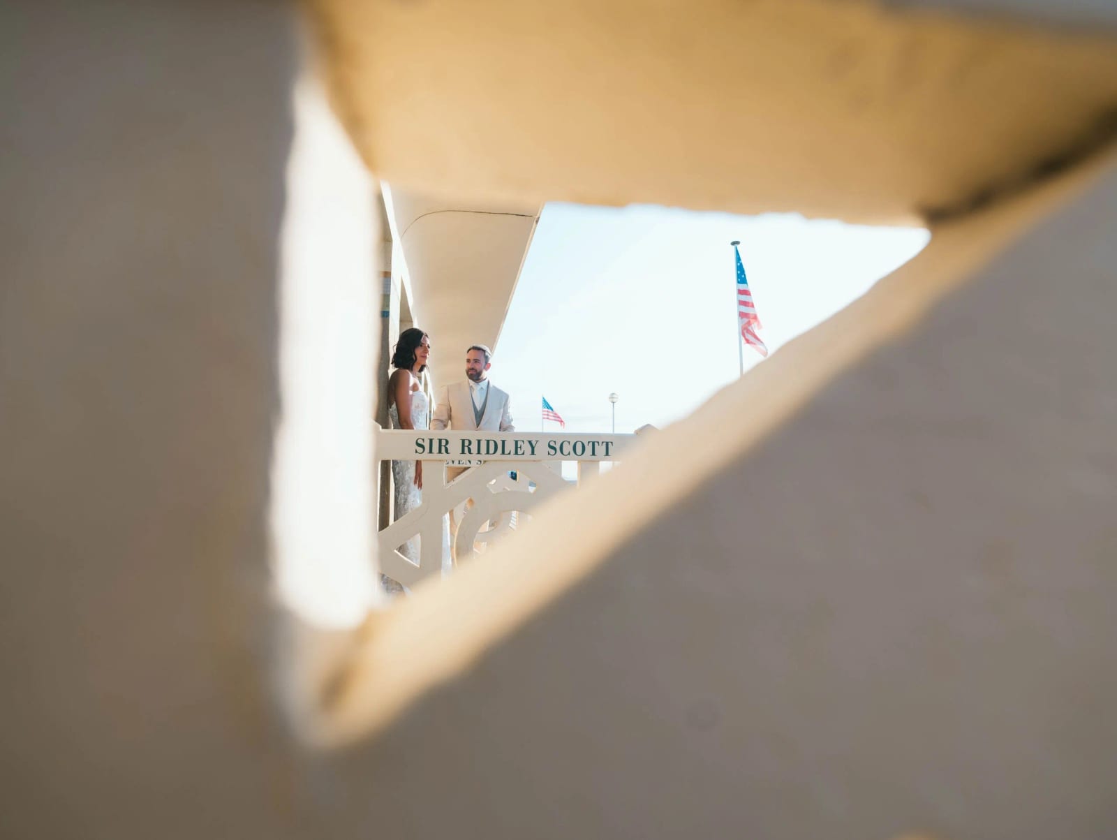 Les mariés devant la cabine Sir Ridley Scott des Planches de Deauville, cadrés à travers un ajour de béton, drapeau américain, mariage au Pavillon des Bains, Deauville, par Jean-Baptiste Argyre (Jaysphotography)