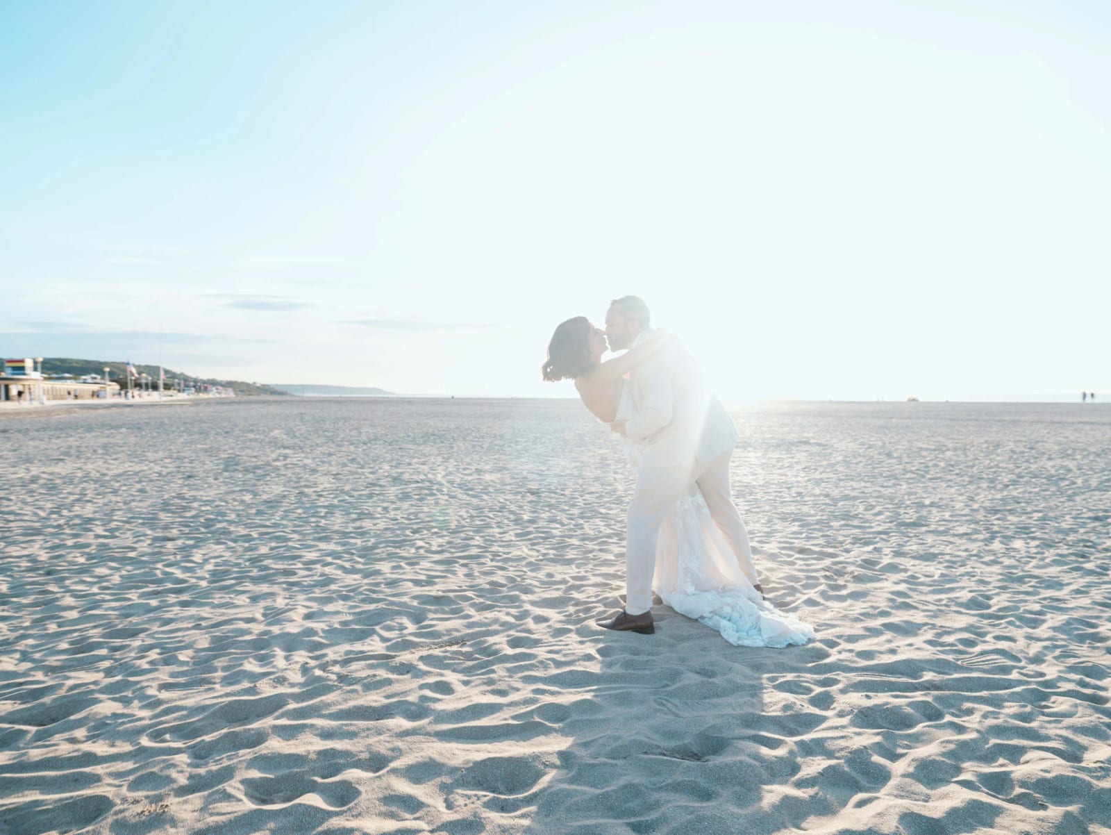 Baiser renversé des mariés à contre-jour sur le sable de la plage de Deauville, front de mer au loin, mariage au Pavillon des Bains, Deauville, par Jean-Baptiste Argyre (Jaysphotography)