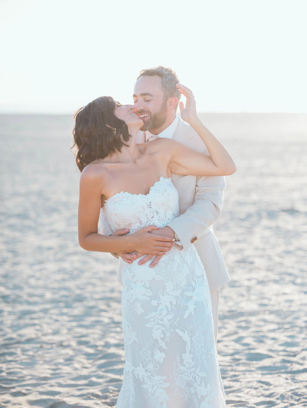 Le marié embrasse la mariée dans le cou sur la plage, robe de dentelle sans bretelles, mariage au Pavillon des Bains, Deauville, par Jean-Baptiste Argyre (Jaysphotography)