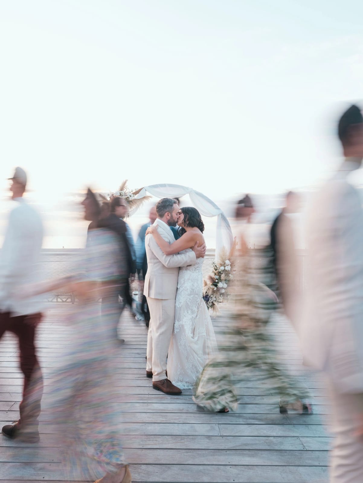 Baiser des mariés sous l'arche, invités en mouvement flous autour d'eux sur la terrasse, mariage au Pavillon des Bains, Deauville, par Jean-Baptiste Argyre (Jaysphotography)