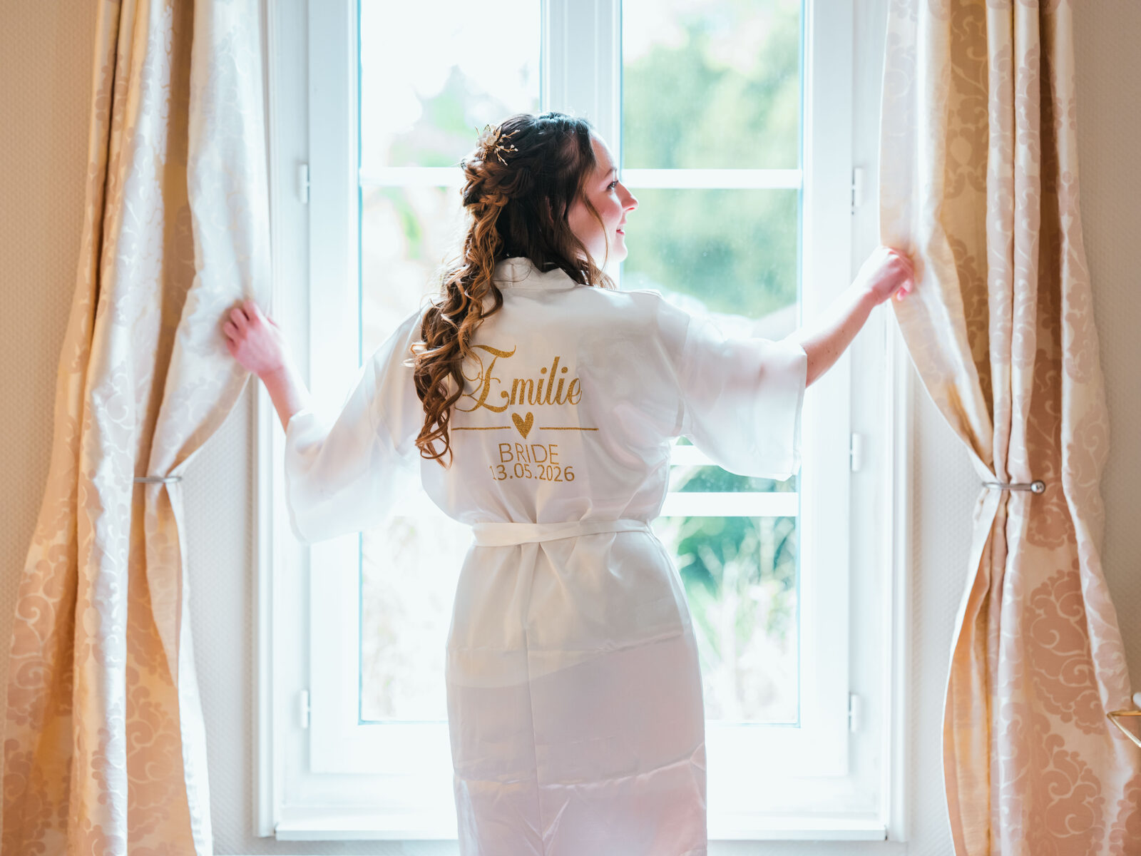 Émilie de dos en peignoir personnalisé Bride 13.05.2026 ouvre les rideaux de sa suite pendant les préparatifs — mariage au Château de Naours, par Jean-Baptiste Argyre (Jaysphotography)