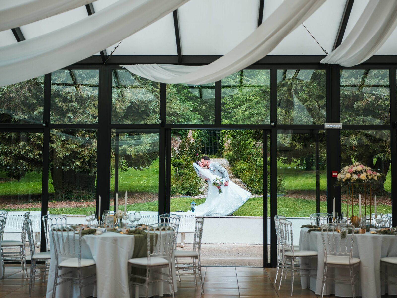 Les mariés s'embrassent dans le parc, vus à travers la verrière de la salle de réception dressée — mariage au Château de Naours, par Jean-Baptiste Argyre (Jaysphotography)