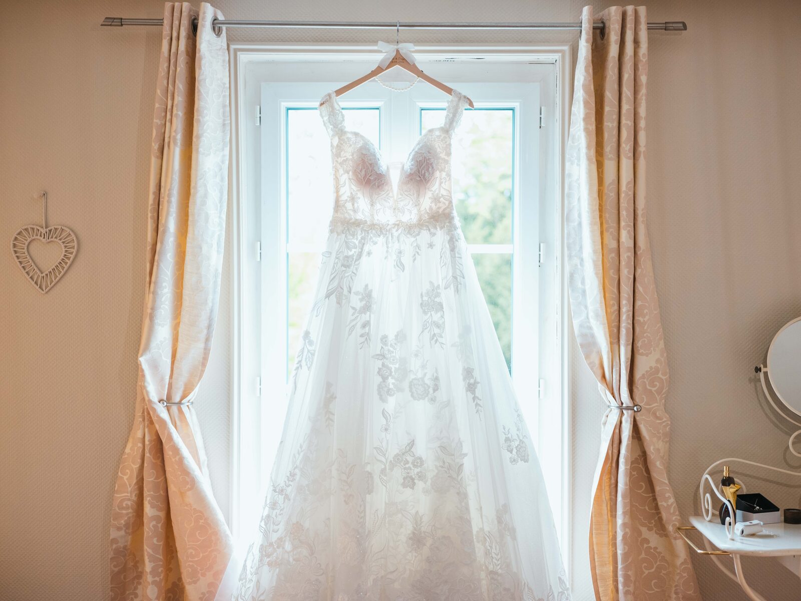 La robe de mariée en dentelle florale d'Émilie suspendue devant la fenêtre de la suite — mariage au Château de Naours, par Jean-Baptiste Argyre (Jaysphotography)