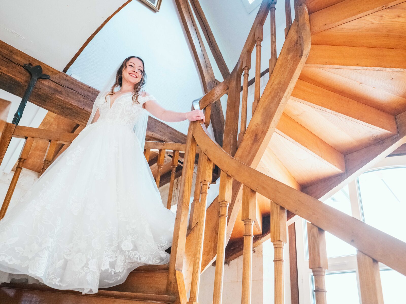 Émilie descend l'escalier en colimaçon en bois du château, robe à traîne et grand sourire — mariage au Château de Naours, par Jean-Baptiste Argyre (Jaysphotography)