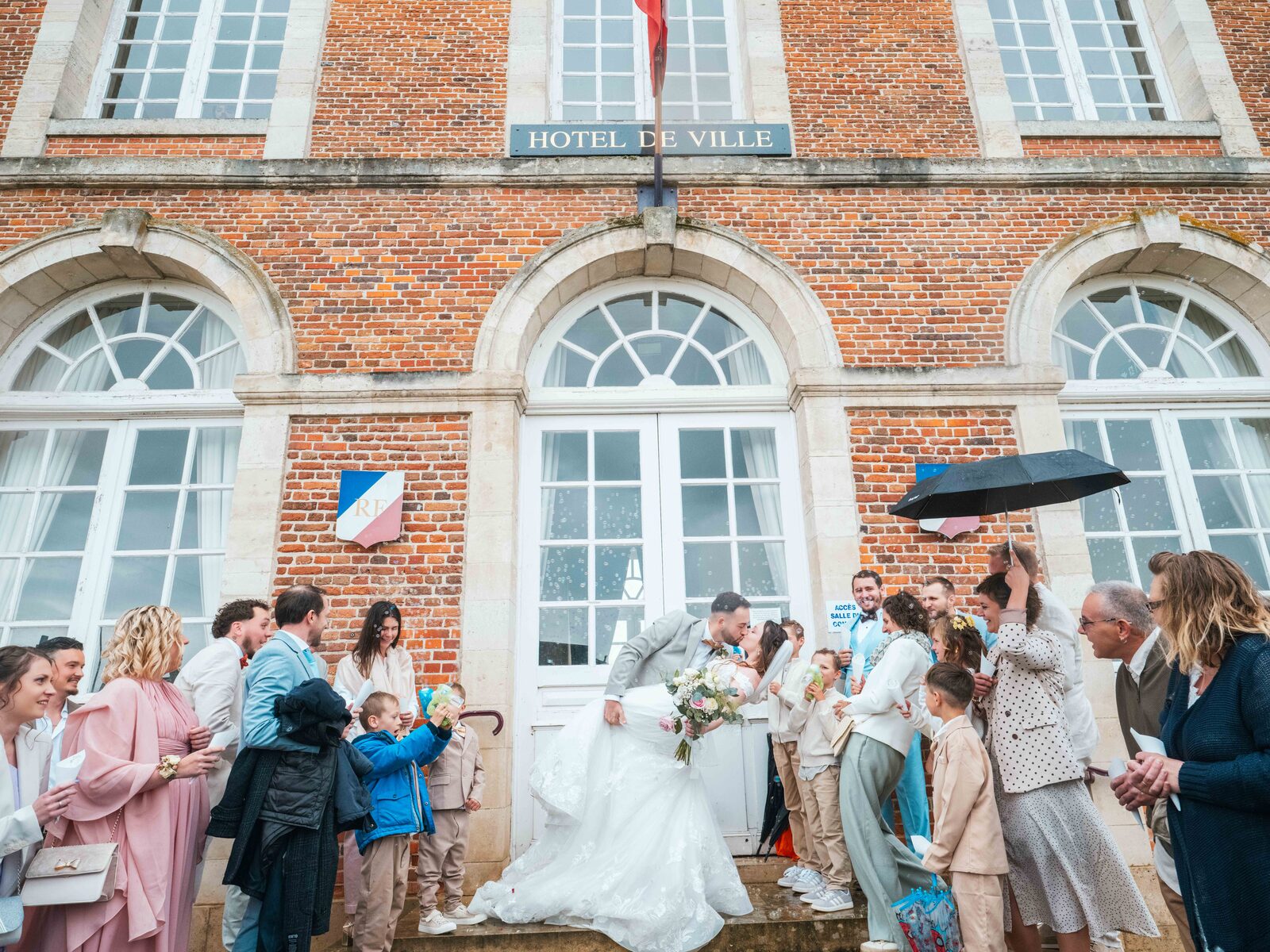 Sortie sous l'averse devant l'Hôtel de Ville de Bresles, invités et parapluies ouverts — mariage au Château de Naours, par Jean-Baptiste Argyre (Jaysphotography)