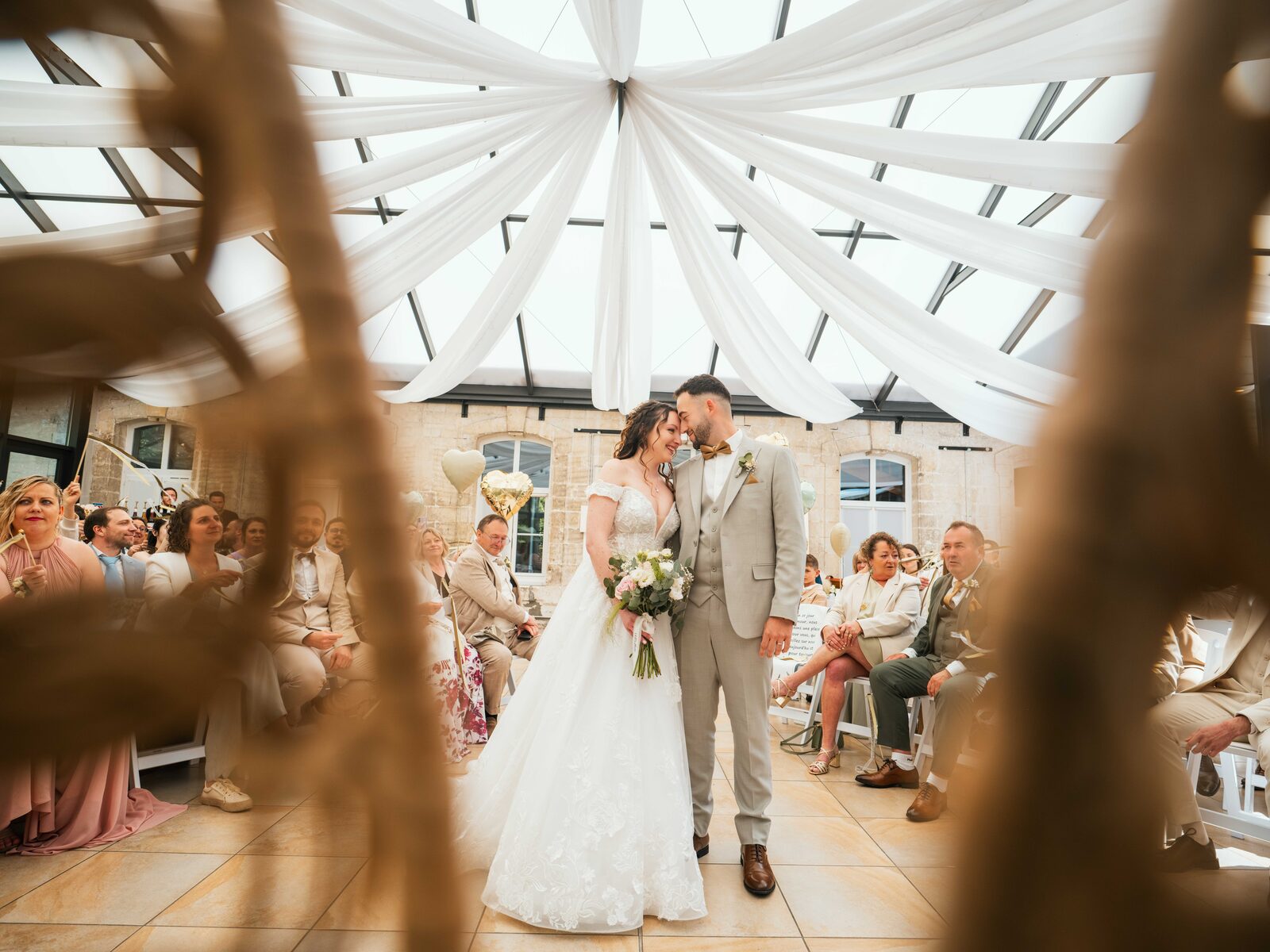 Baiser des mariés dans l'allée centrale sous la verrière, invités assis, cérémonie laïque — mariage au Château de Naours, par Jean-Baptiste Argyre (Jaysphotography)