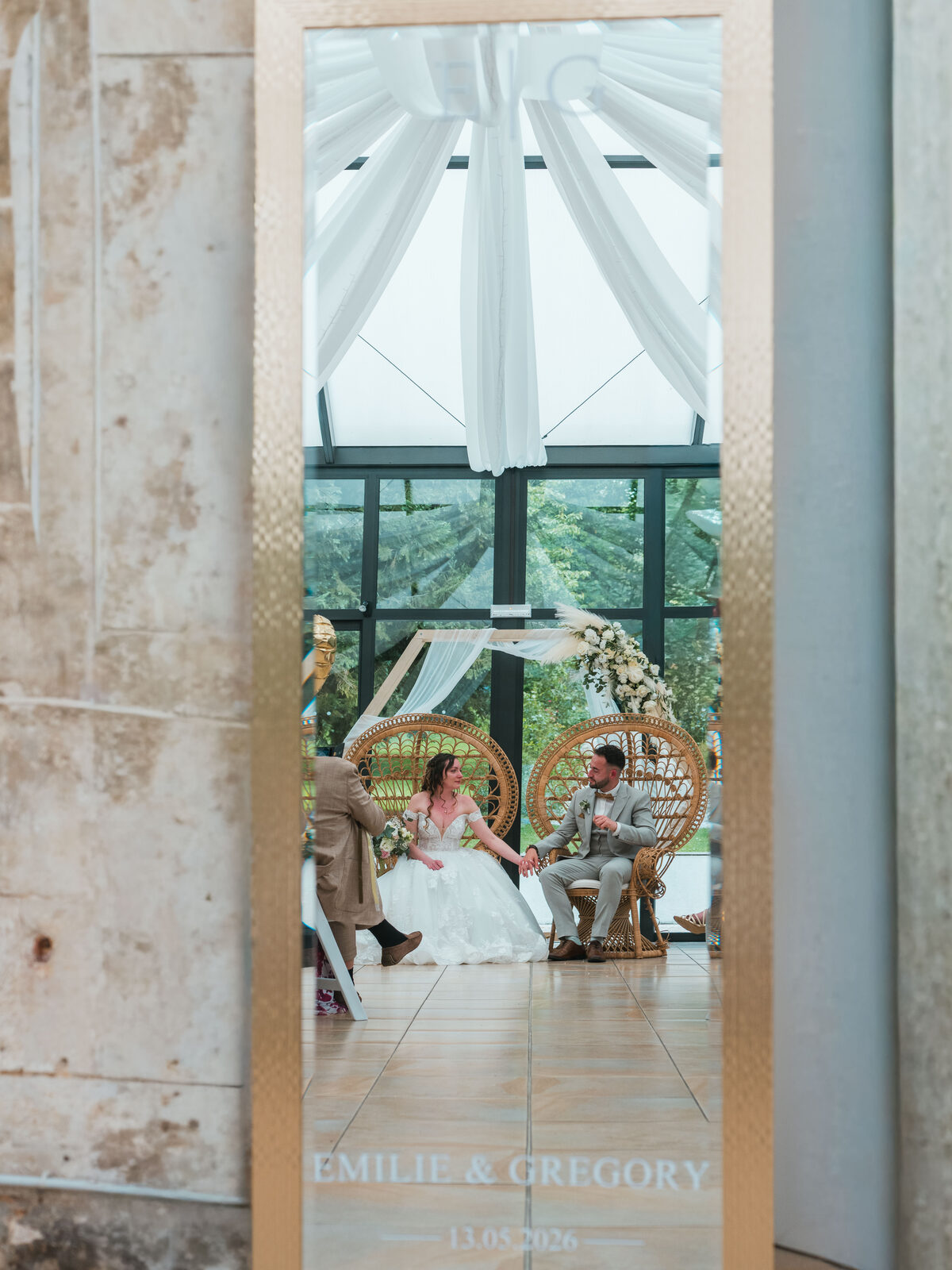 Reflet dans le miroir doré Émilie & Grégory : les mariés main dans la main sur leurs fauteuils en rotin — mariage au Château de Naours, par Jean-Baptiste Argyre (Jaysphotography)