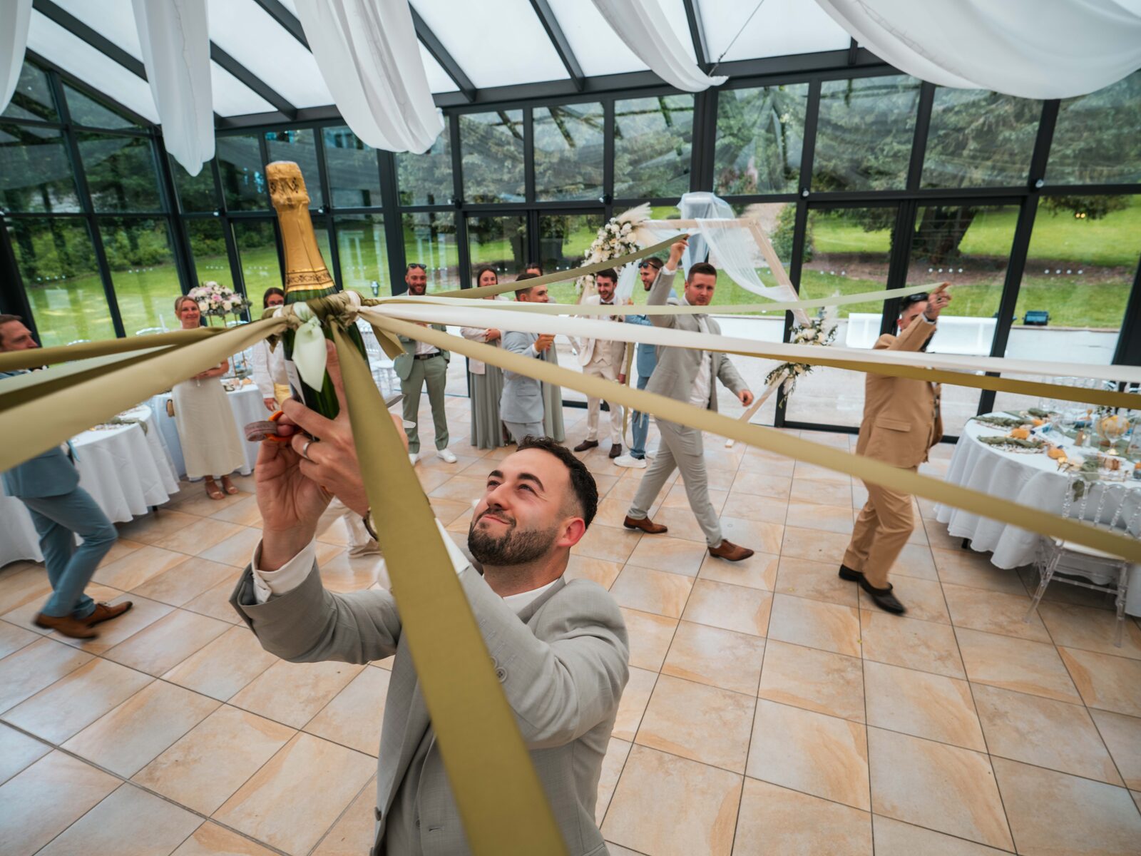 Grégory brandit la bouteille de champagne du jeu du ruban tenue par les invités — mariage au Château de Naours, par Jean-Baptiste Argyre (Jaysphotography)