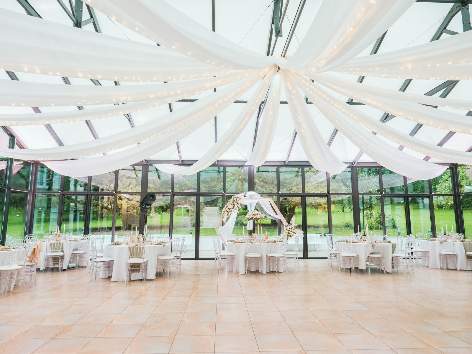 Salle de réception sous la verrière, guirlandes lumineuses et tables rondes dressées — mariage au Château de Naours, par Jean-Baptiste Argyre (Jaysphotography)