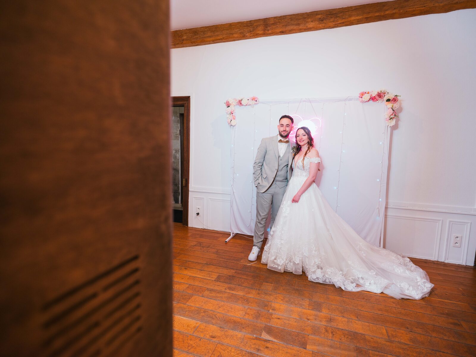 Les mariés posent devant le fond photo au cœur néon dans un salon du château — mariage au Château de Naours, par Jean-Baptiste Argyre (Jaysphotography)