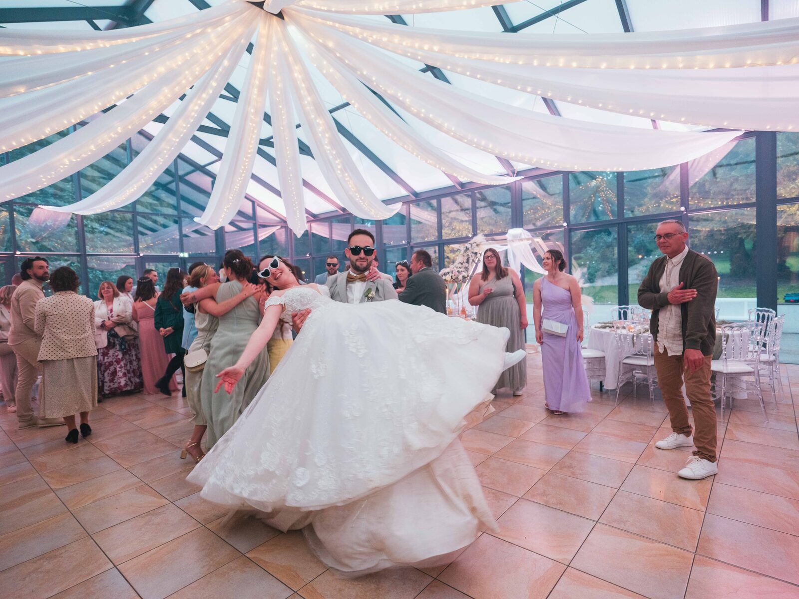 Émilie tournoie dans les bras de Grégory sur la piste, lunettes en cœur, invités autour — mariage au Château de Naours, par Jean-Baptiste Argyre (Jaysphotography)