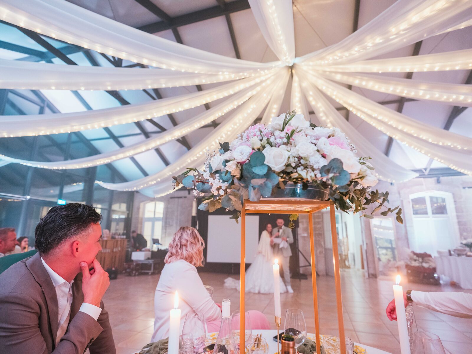 Détail de réception : centre floral et bougies, Grégory observe, les mariés en arrière-plan — mariage au Château de Naours, par Jean-Baptiste Argyre (Jaysphotography)