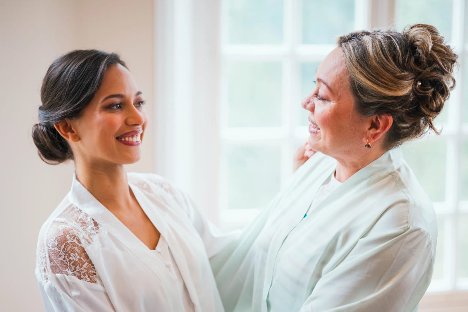 Ana, chignon bas et peignoir en dentelle, face à une proche qui ajuste son collier pendant la mise en beauté, devant une fenêtre lumineuse du château du Domaine d'Orgival, mariage au Domaine d'Orgival, par Jean-Baptiste Argyre (Jaysphotography)