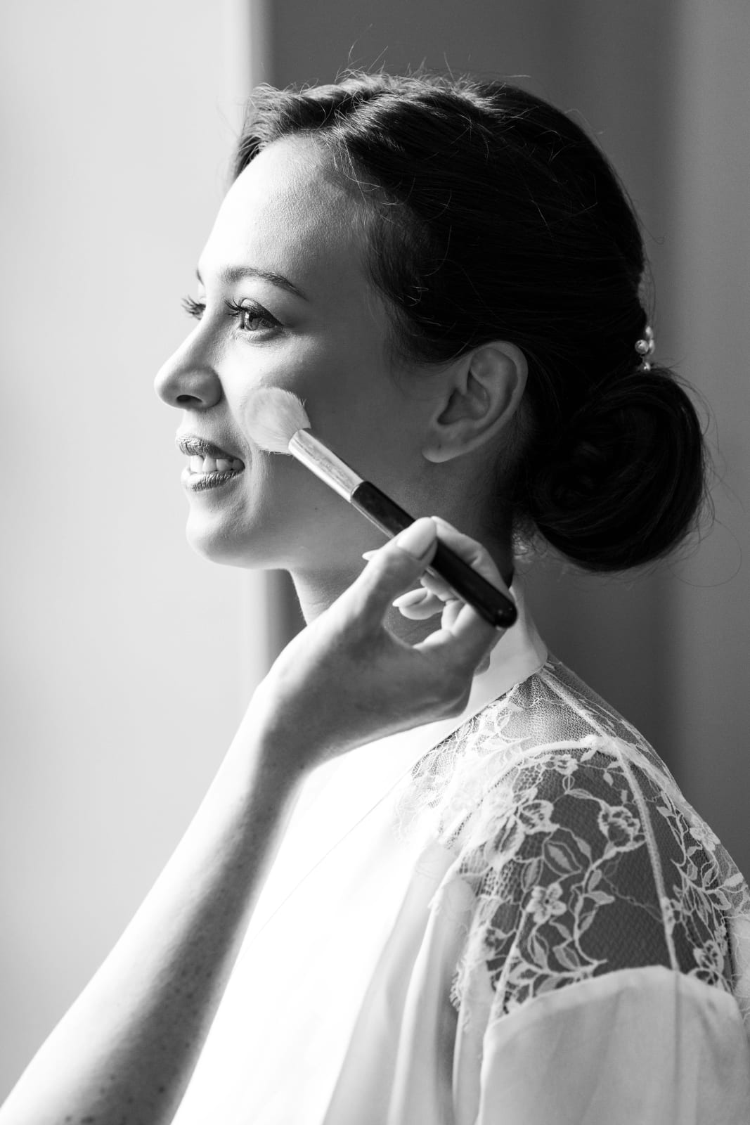 Portrait en noir et blanc d'Ana de profil pendant son maquillage, une maquilleuse passant le pinceau de teint sur sa joue, mariage au Domaine d'Orgival, par Jean-Baptiste Argyre (Jaysphotography)